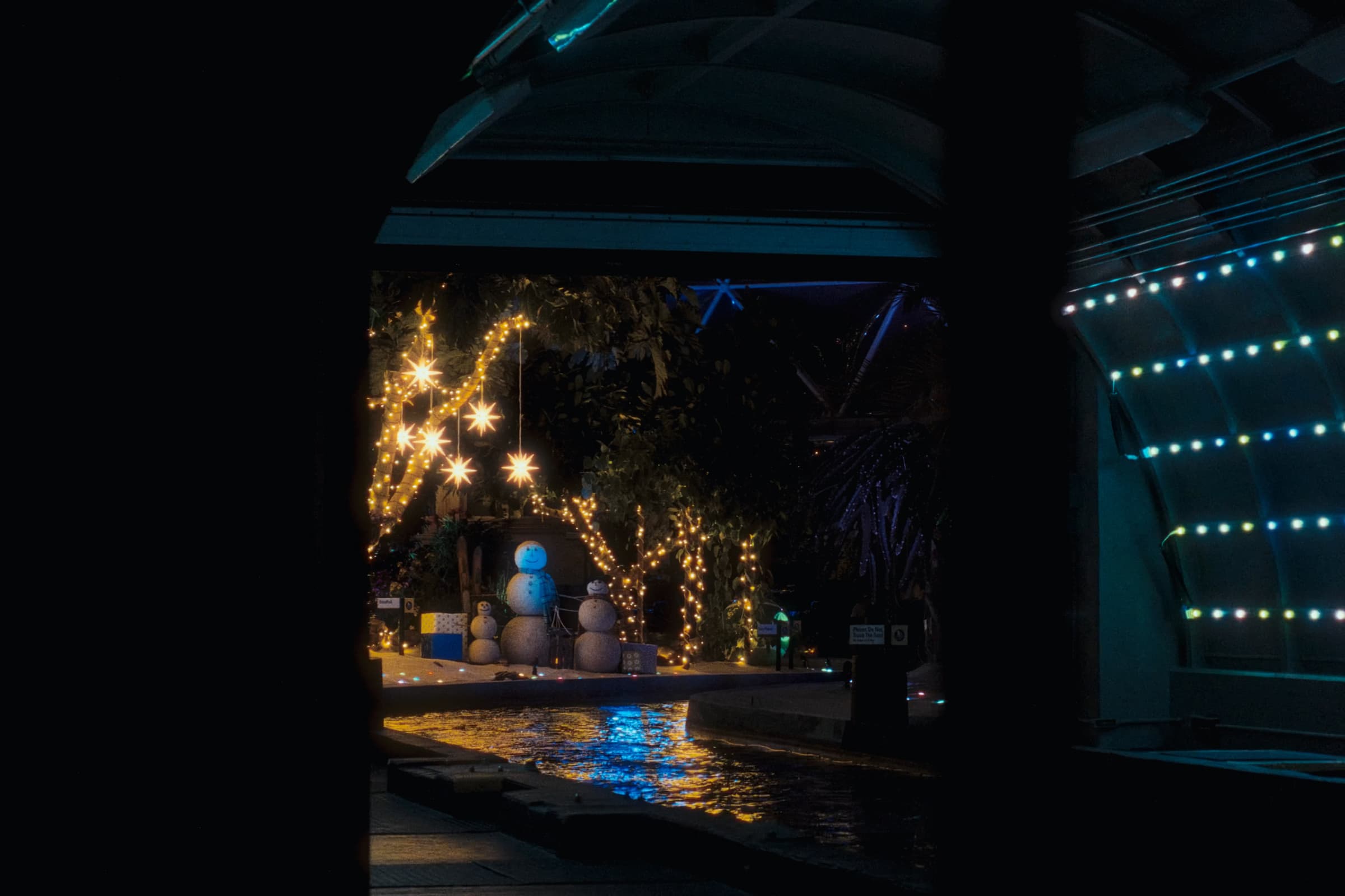 Holiday lights and snowmen beside a water channel under night lighting.