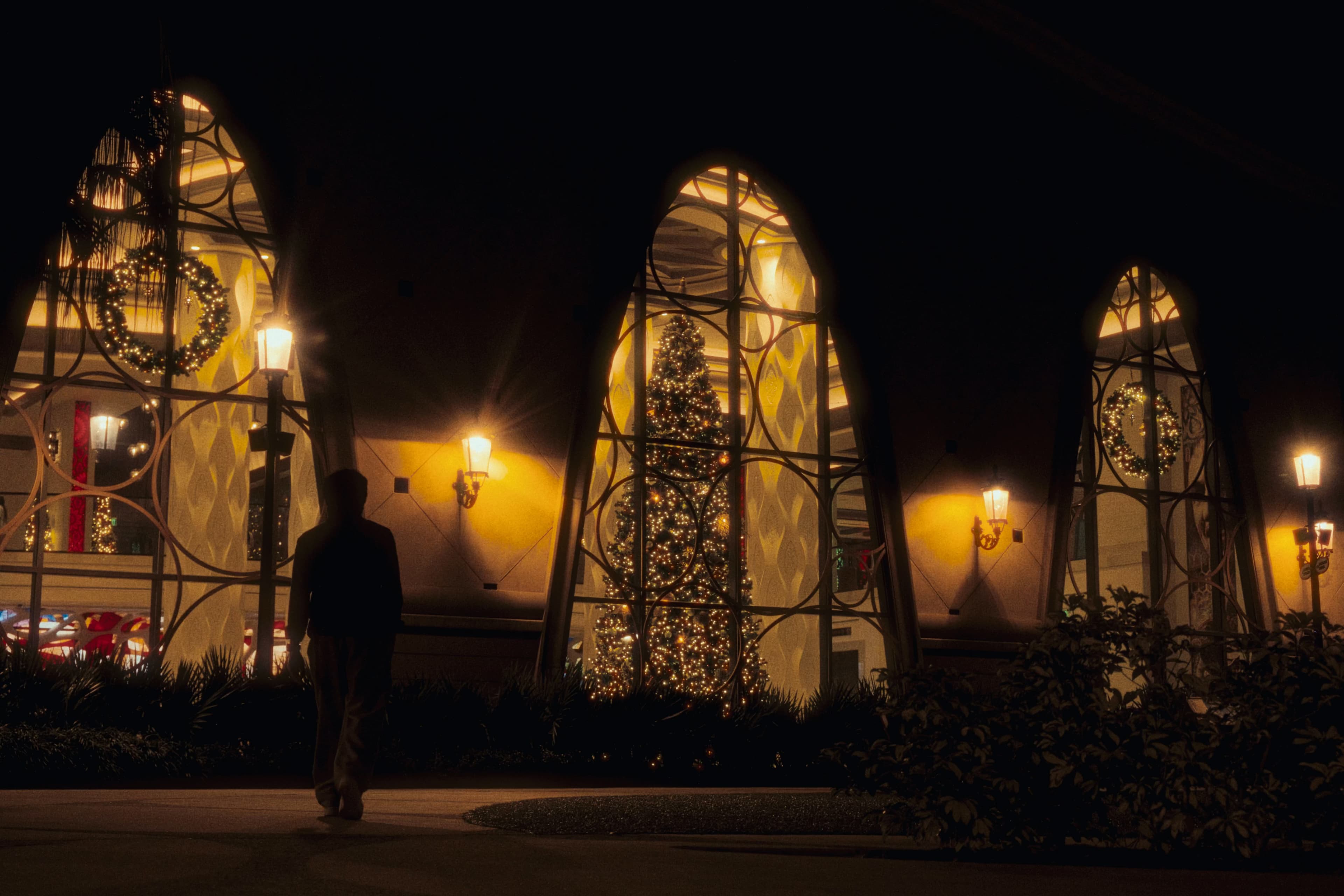 A silhouette walks past arched windows glowing with Christmas trees and wreaths.