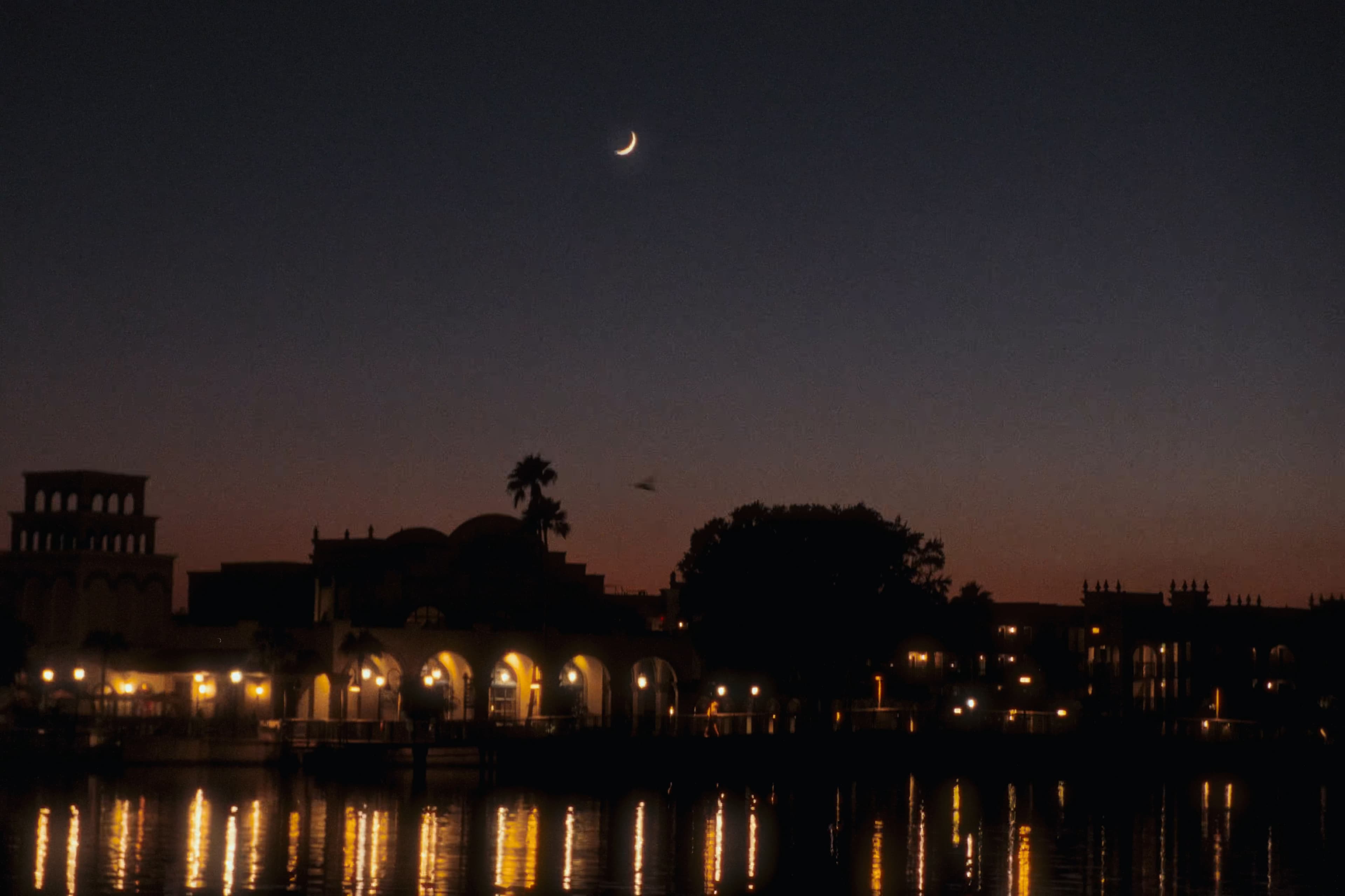 A crescent moon above a waterfront resort with warm reflections on the water.