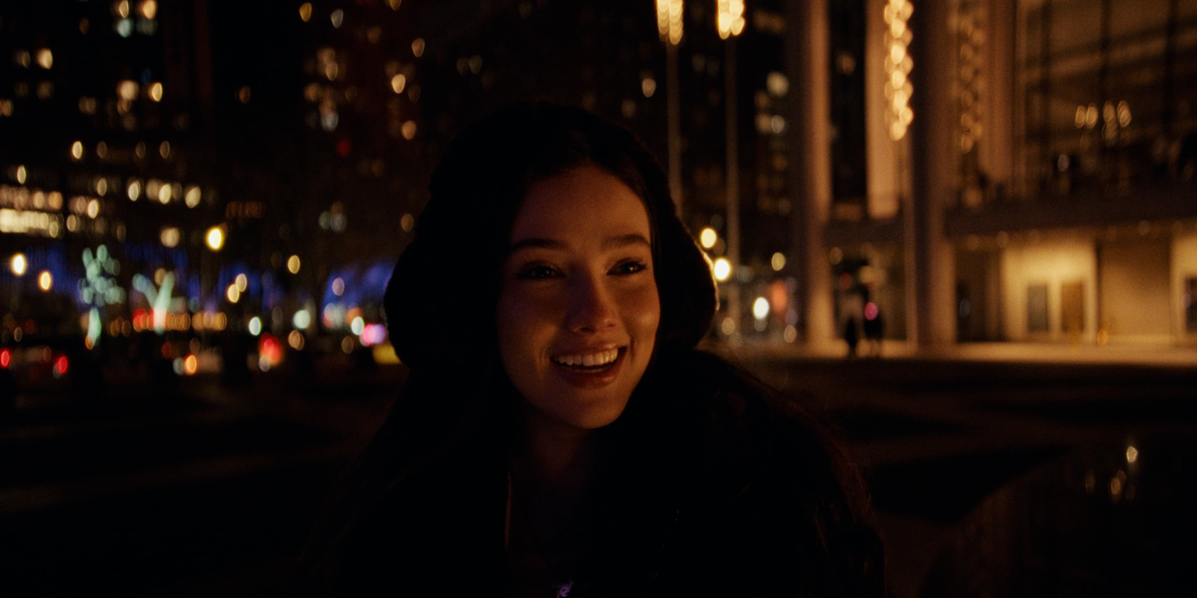 Juliana smiles at night with city lights and buildings softly blurred behind her.