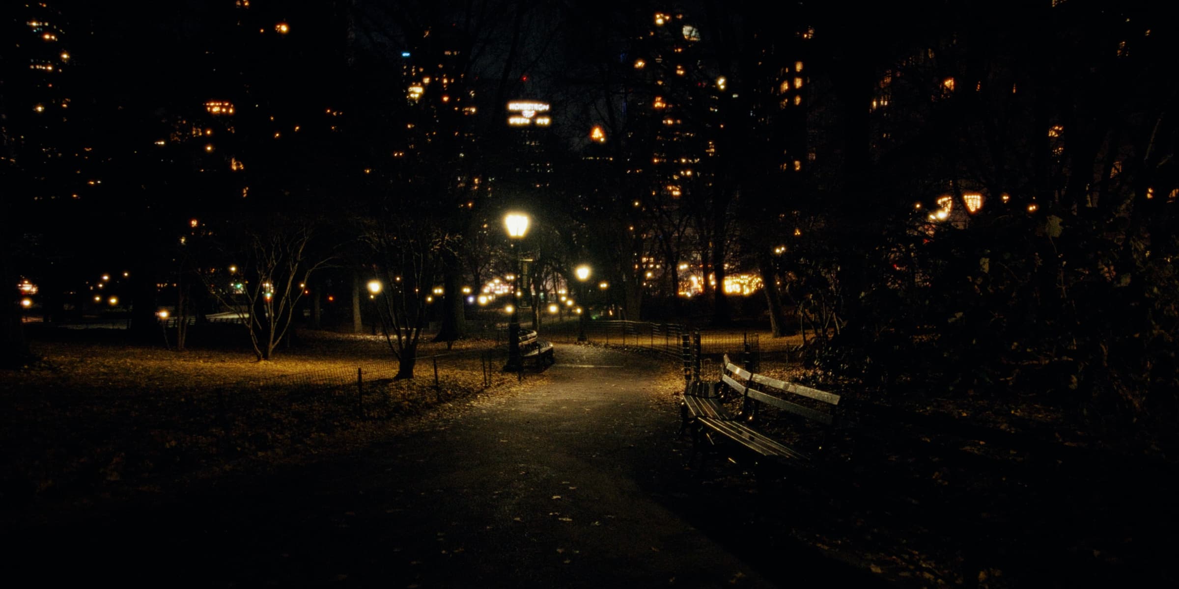 A quiet park path at night with benches and lampposts under city lights.