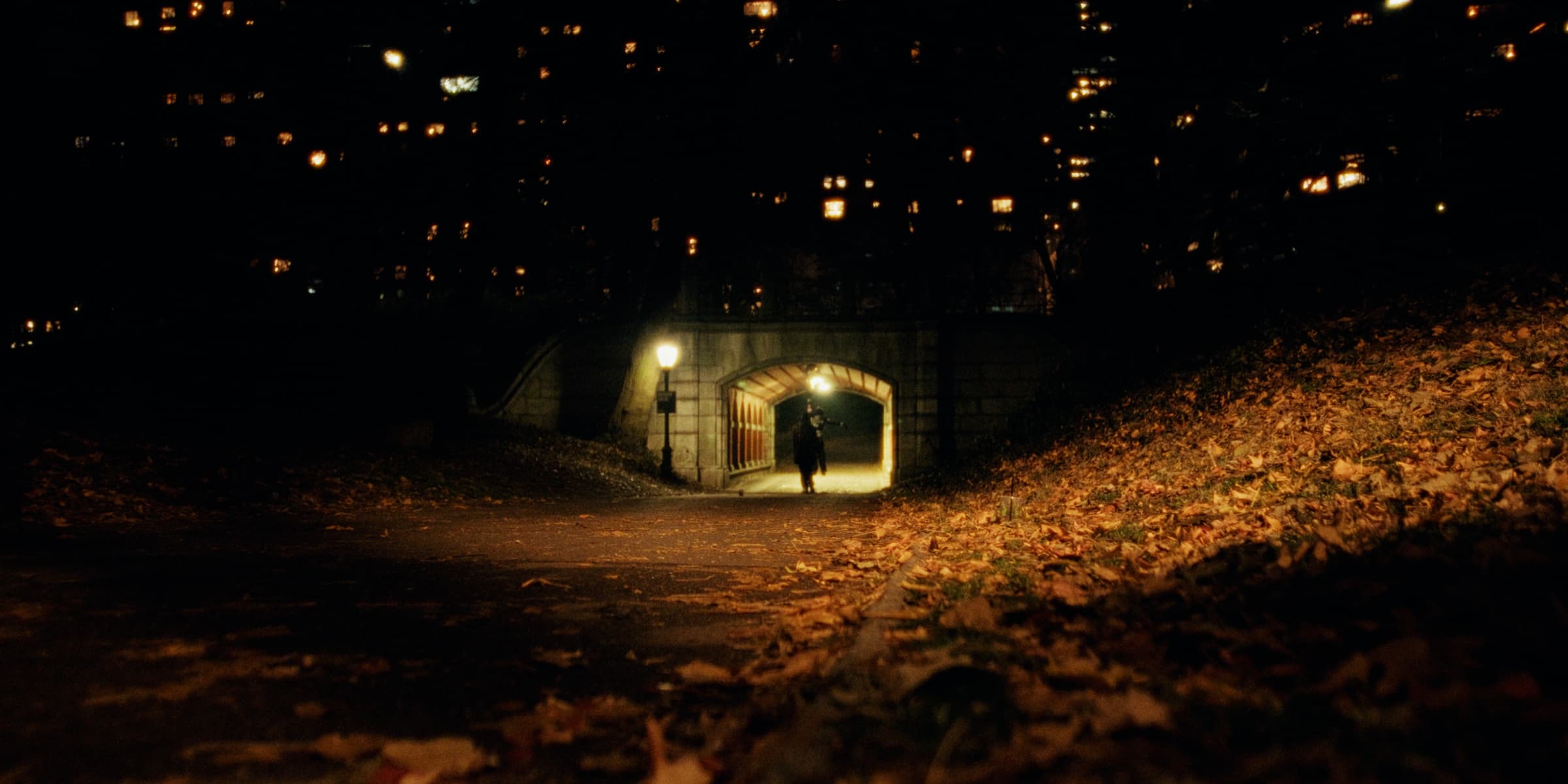 A lone figure walks toward a lit underpass with a stone arch at night.