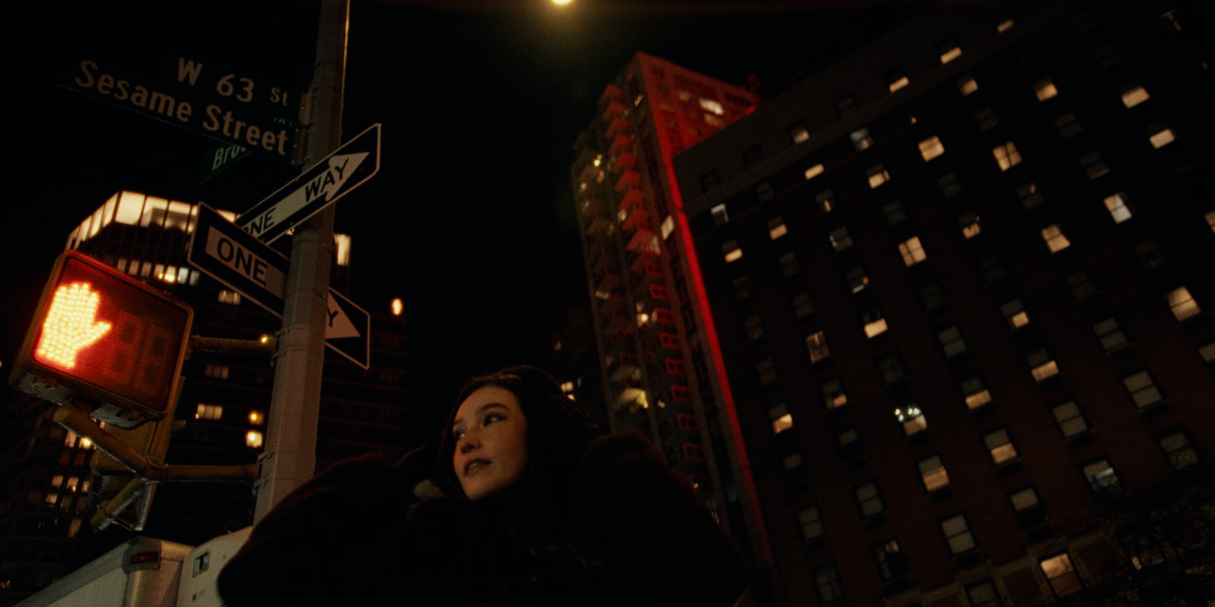 Juliana stands on a corner under a "W 63 St" sign at night with tall buildings behind her.