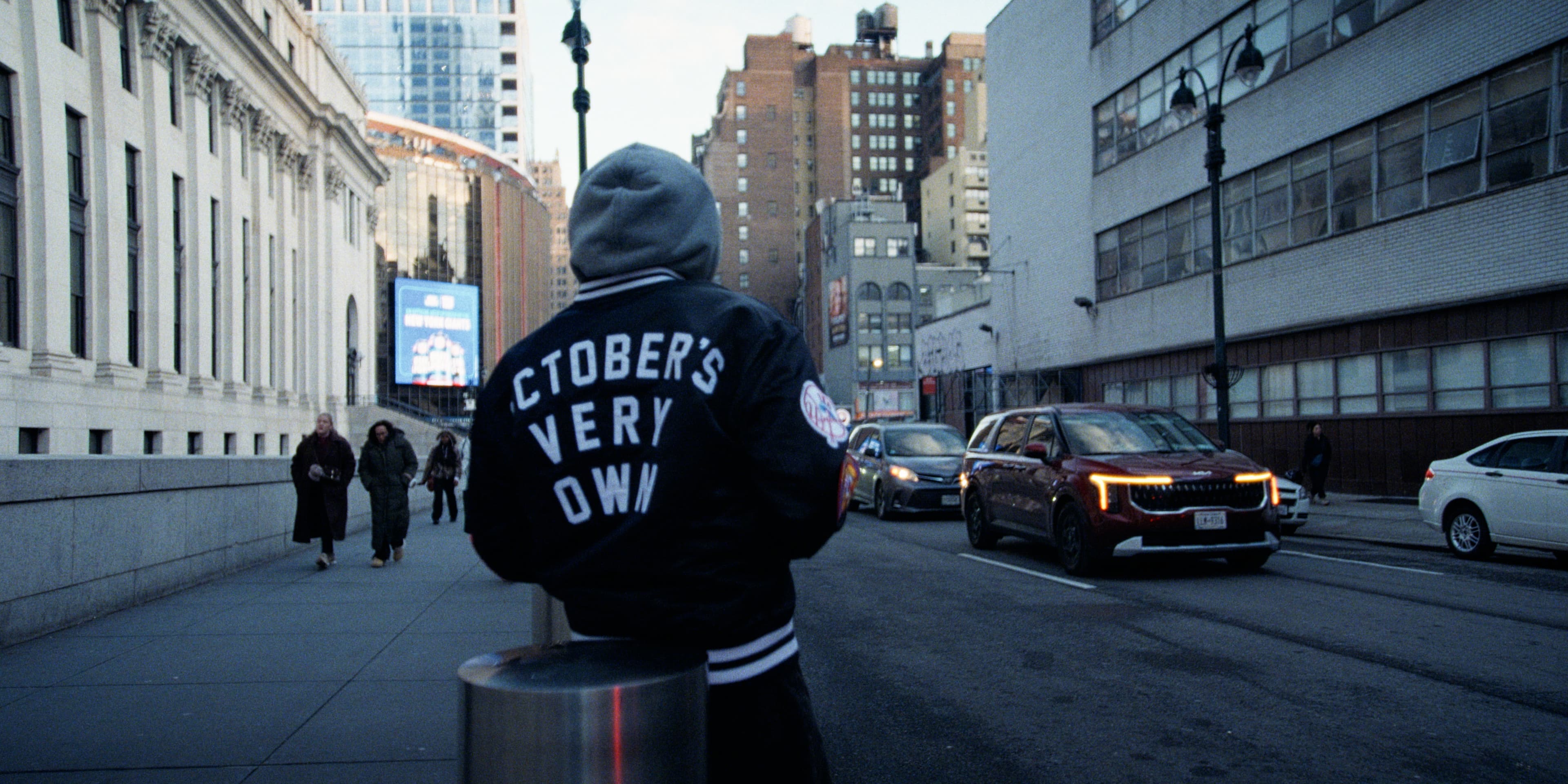 A person in a hoodie walks along a city street in late afternoon light.