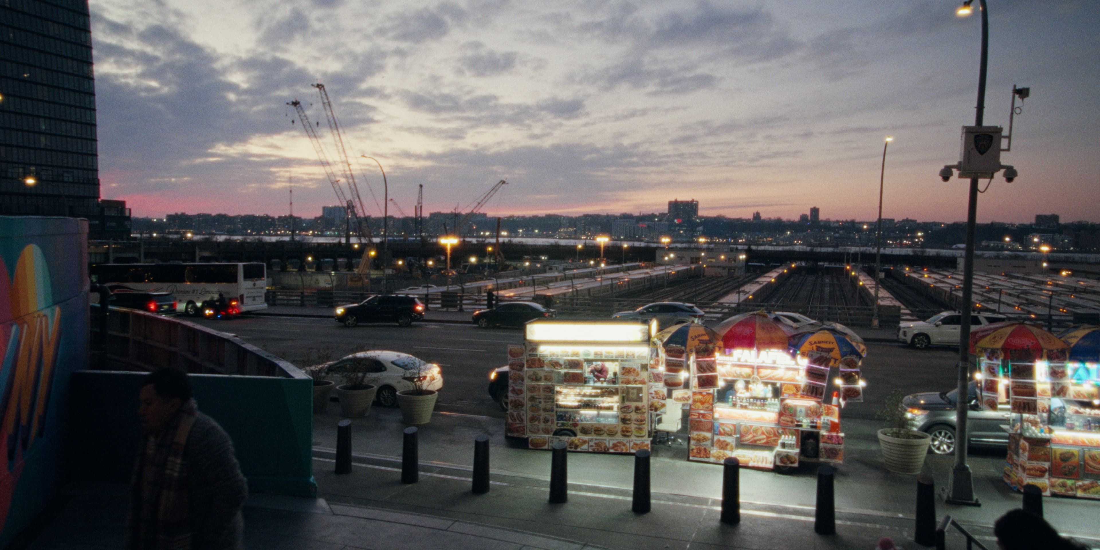 A dusky skyline with cranes and a highway, food carts glowing in the foreground.