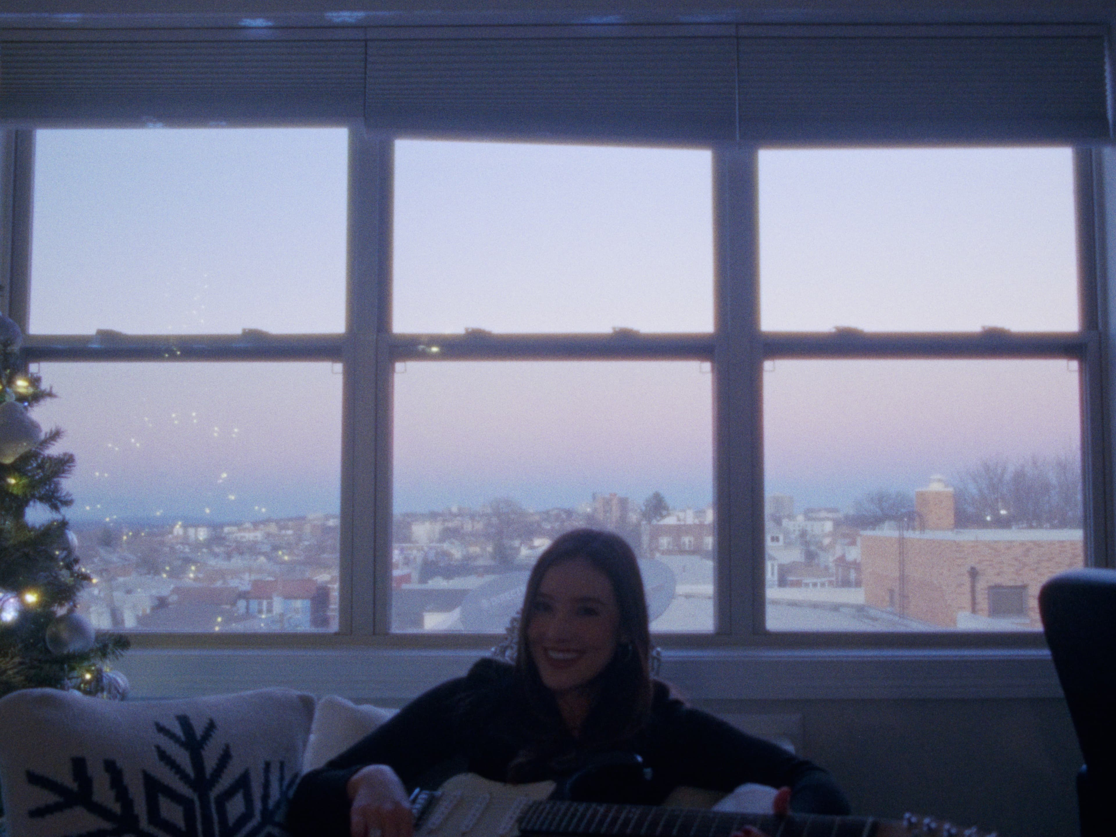 Juliana sits on a couch with a guitar as dusk light fills the windows.