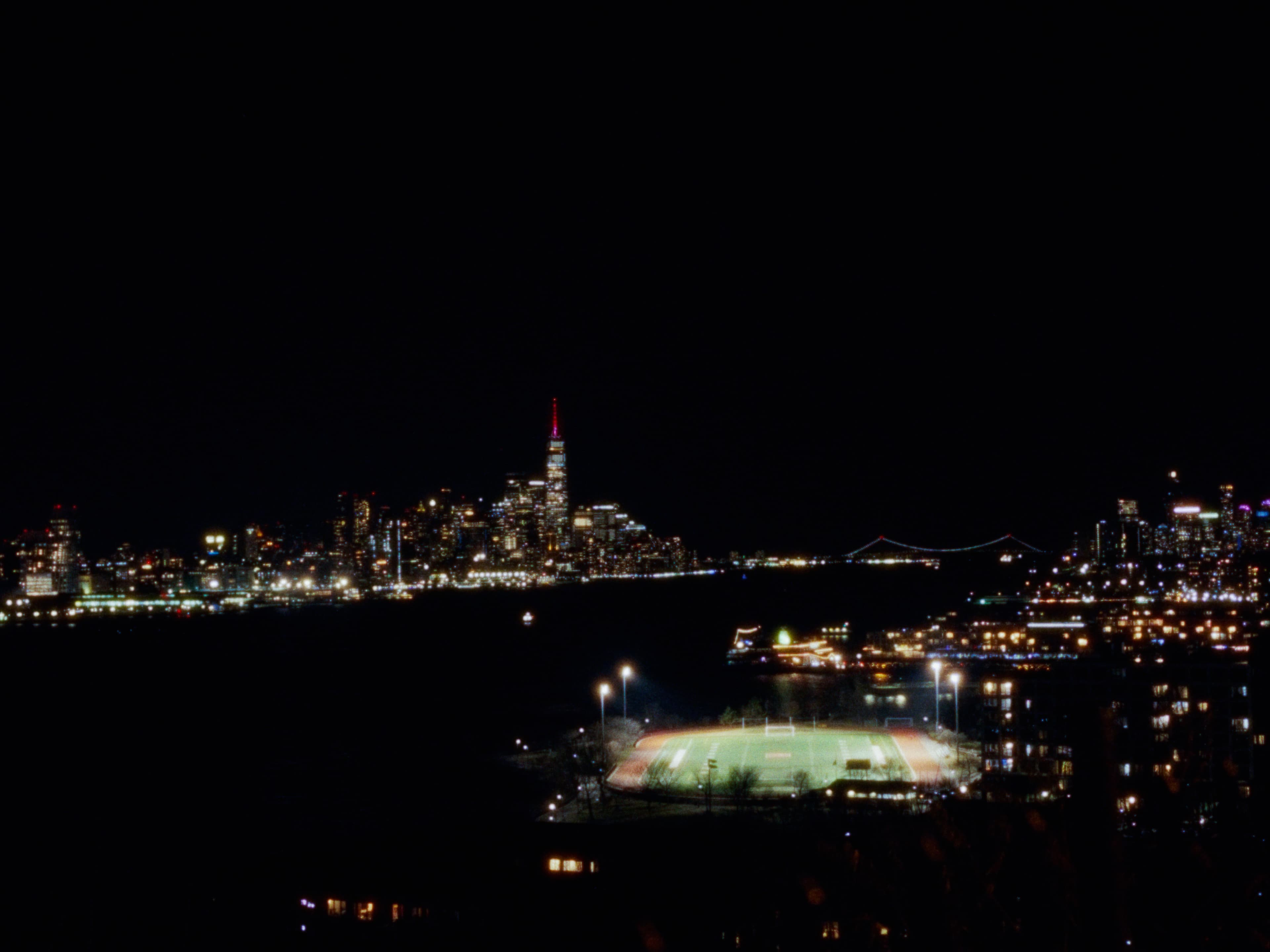A nighttime view of the New York skyline across the water, with a lit sports field in the foreground.