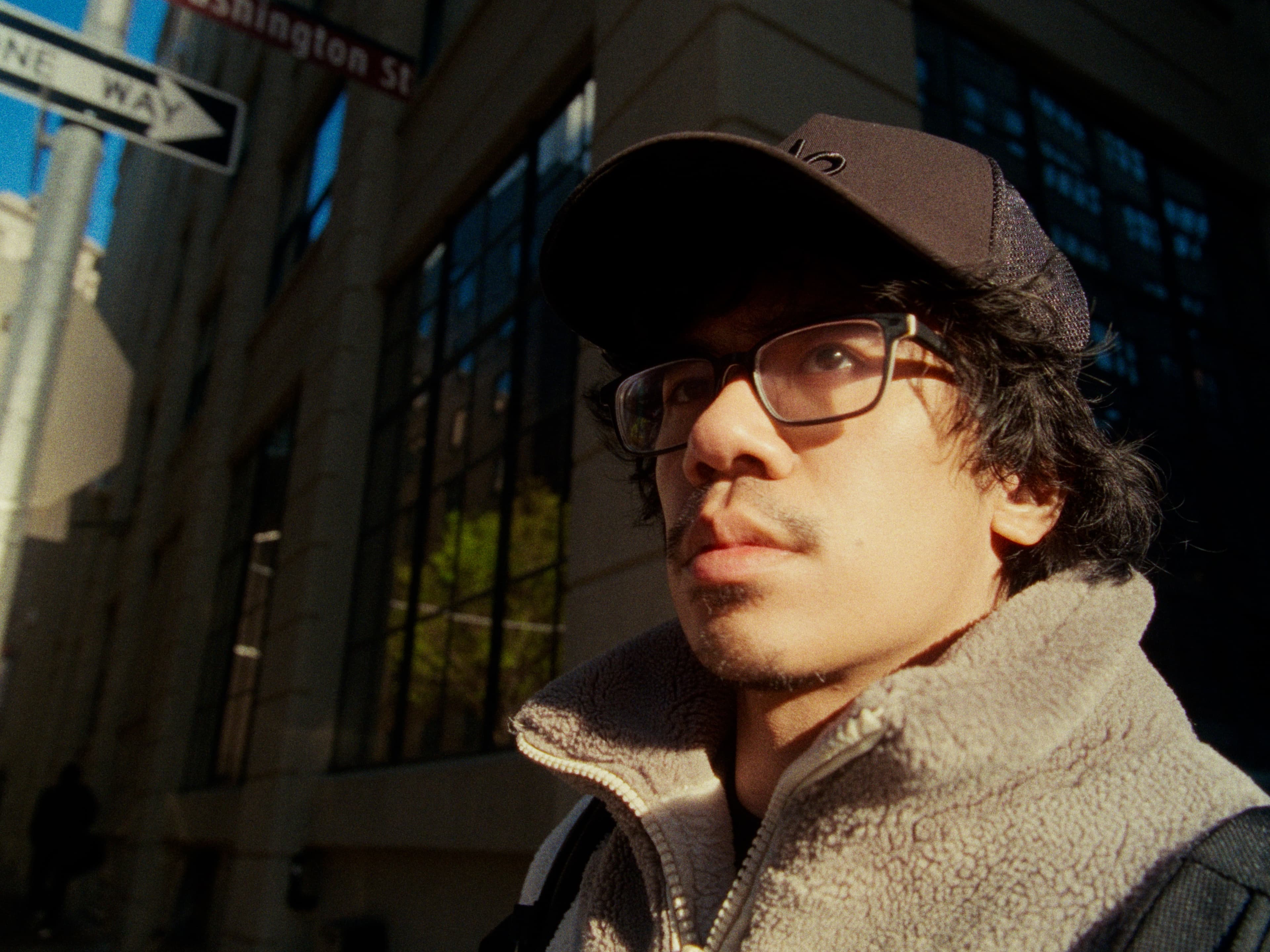 Andrew under a 'Washington St' street sign in cap, glasses, and sherpa fleece — golden afternoon light raking across brick and glass behind.