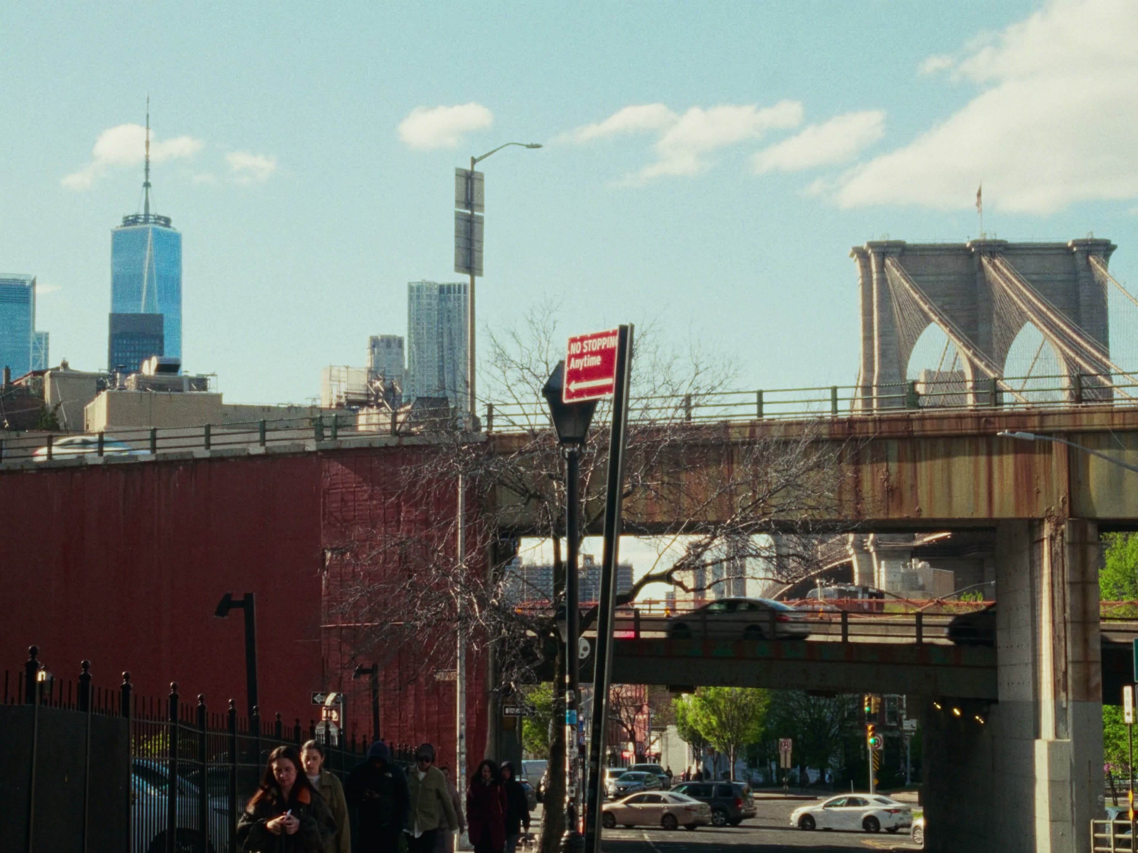A BQE overpass cuts across the frame with a red-brick retaining wall below, the Freedom Tower and Brooklyn Bridge in the distance, a 'NO STOPPING Anytime' sign on a lamppost mid-frame.