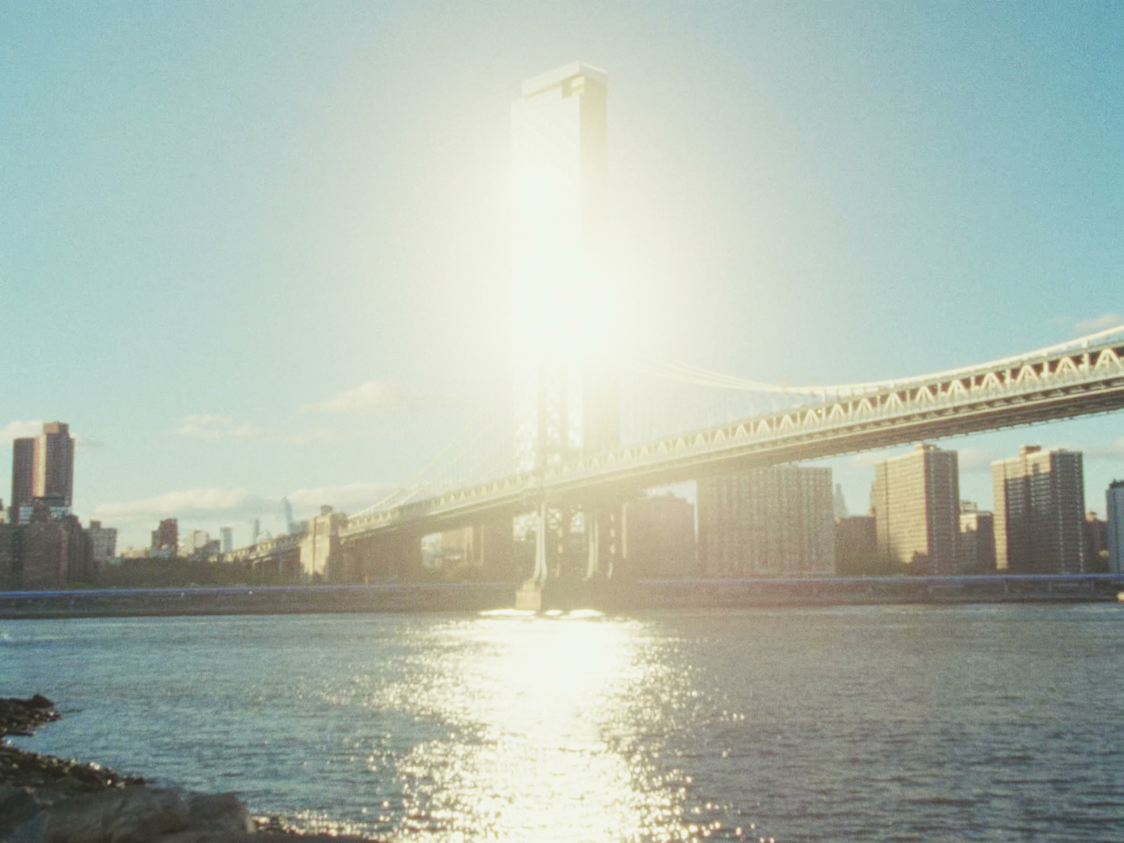 The Manhattan Bridge backlit by a fierce sun — a bright flare blooms behind the main tower, rendering the skyline in soft silhouette.