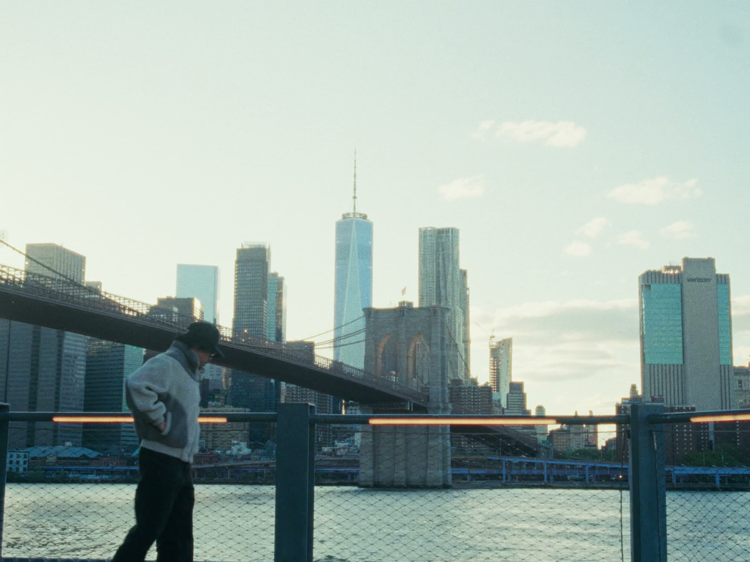 Andrew Amisola in Freezing Cold in Brooklyn — Andrew stands at a chain-link railing in a grey sherpa fleece, the Brooklyn Bridge arch and Lower Manhattan skyline across the water behind him.