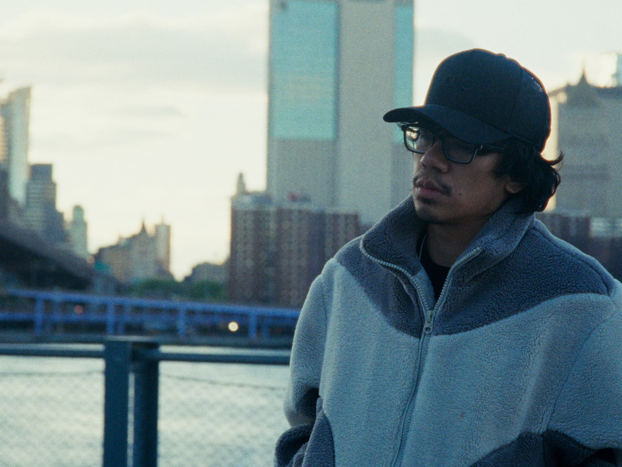Andrew Amisola in Freezing Cold in Brooklyn — Andrew at the pier, zipped into a sherpa fleece against the cold, gaze turned away from the city, Manhattan softening into dusk behind.