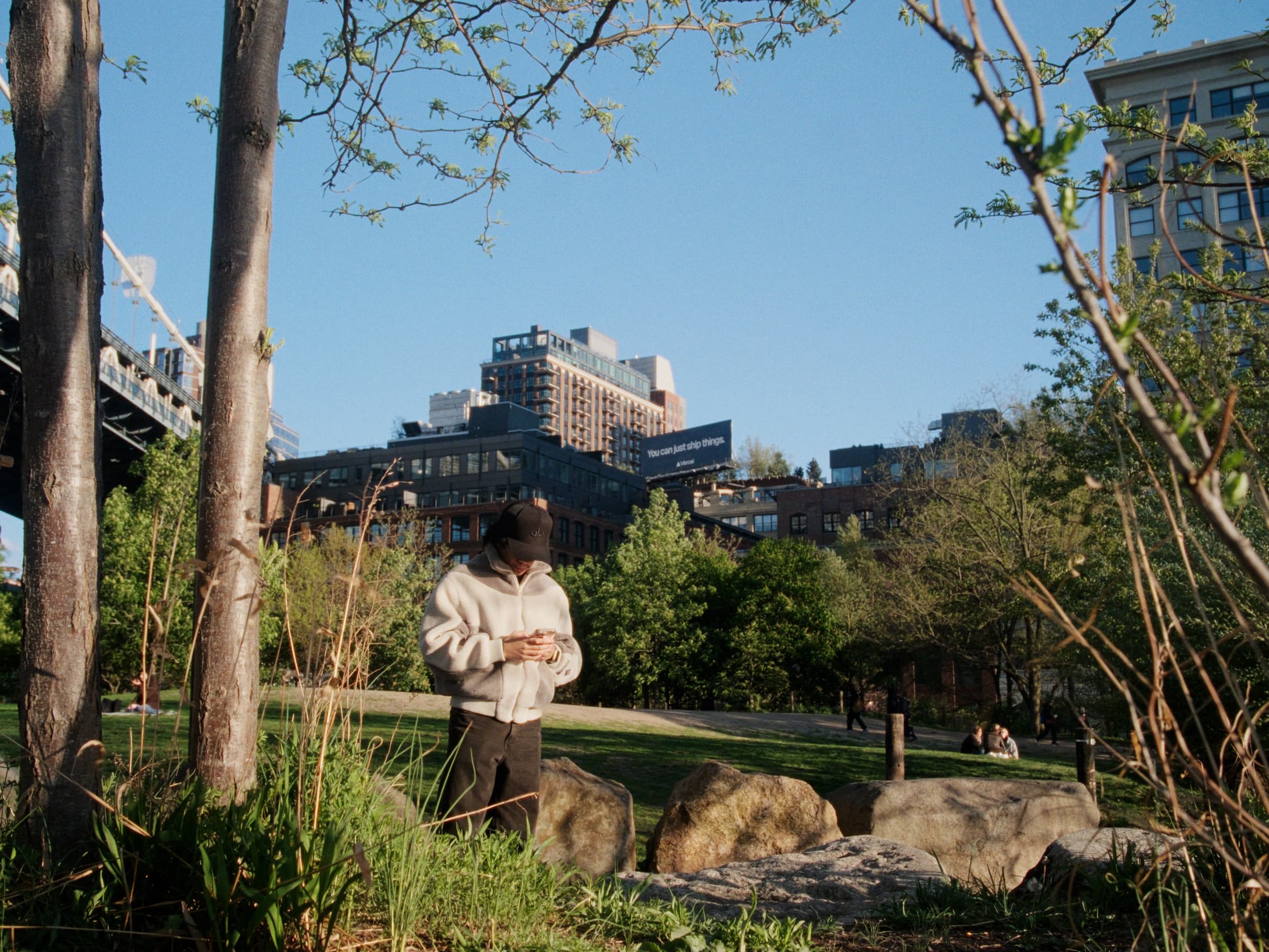 Andrew Amisola in Freezing Cold in Brooklyn — Andrew stands among large boulders in a park clearing in his sherpa fleece, bare trees framing the shot, low residential buildings and sky beyond.