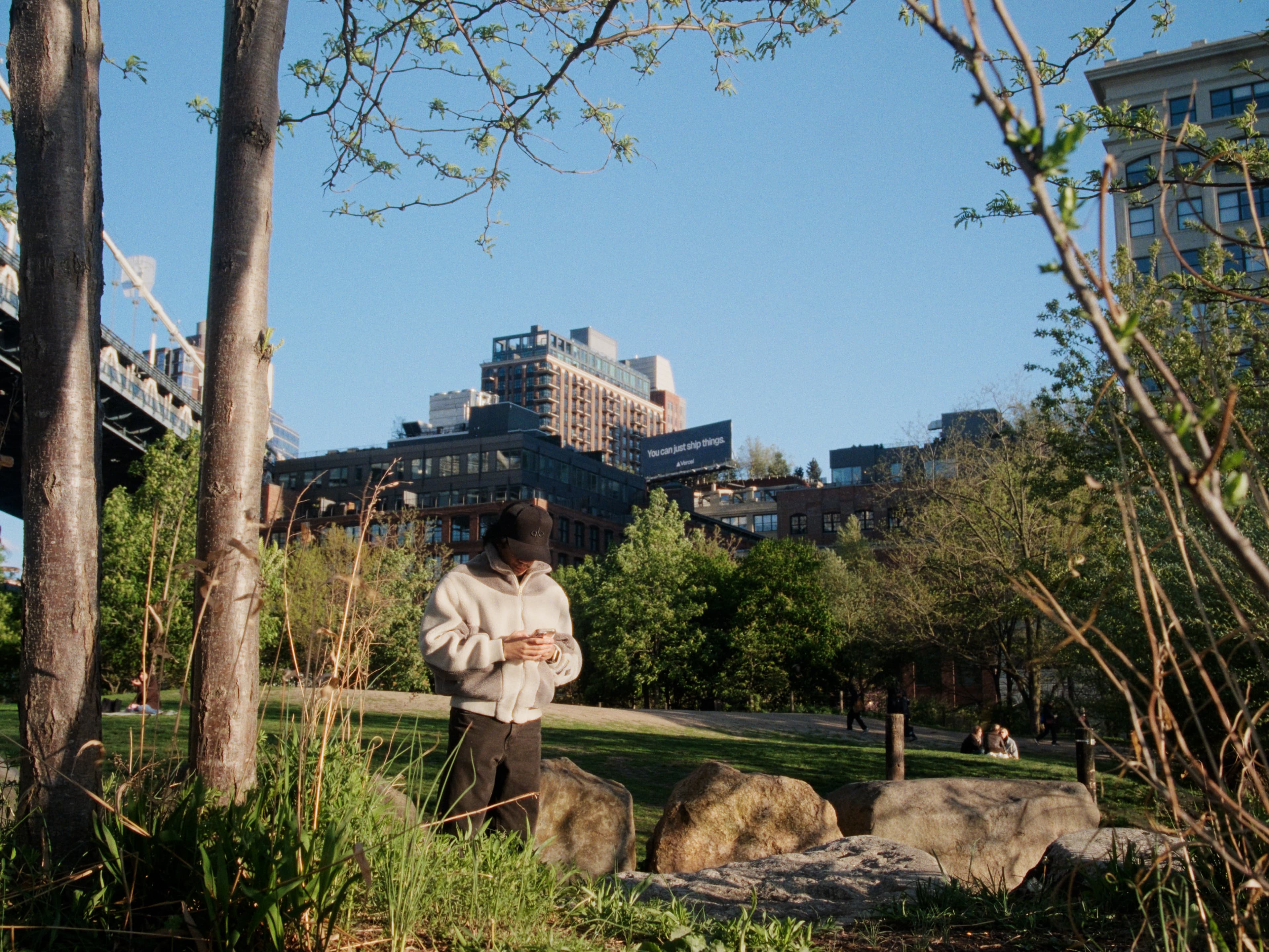 Andrew stands among large boulders in a park clearing in his sherpa fleece, bare trees framing the shot, low residential buildings and sky beyond.