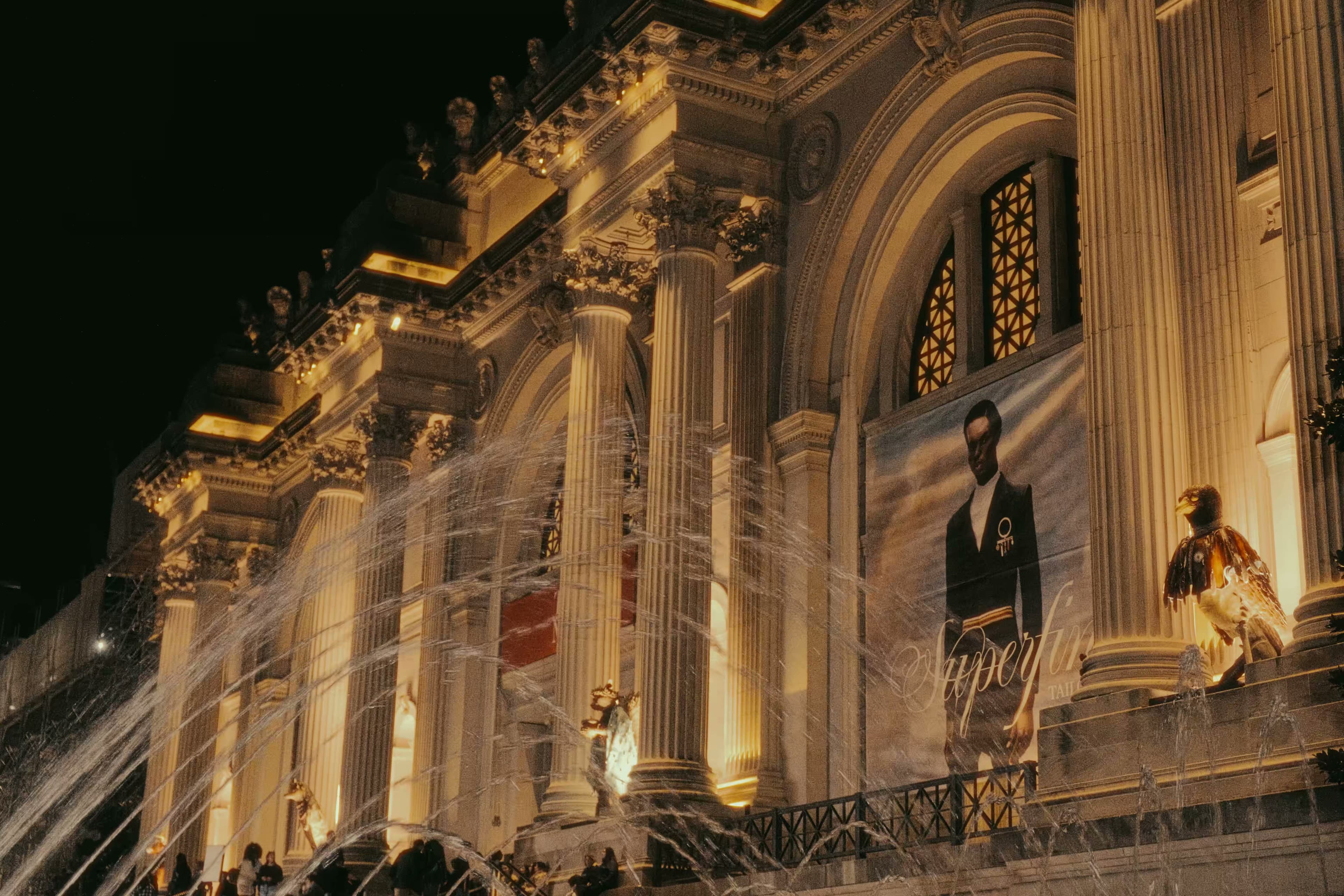 The Met's illuminated Beaux-Arts facade at night with fountains and exhibition banners.