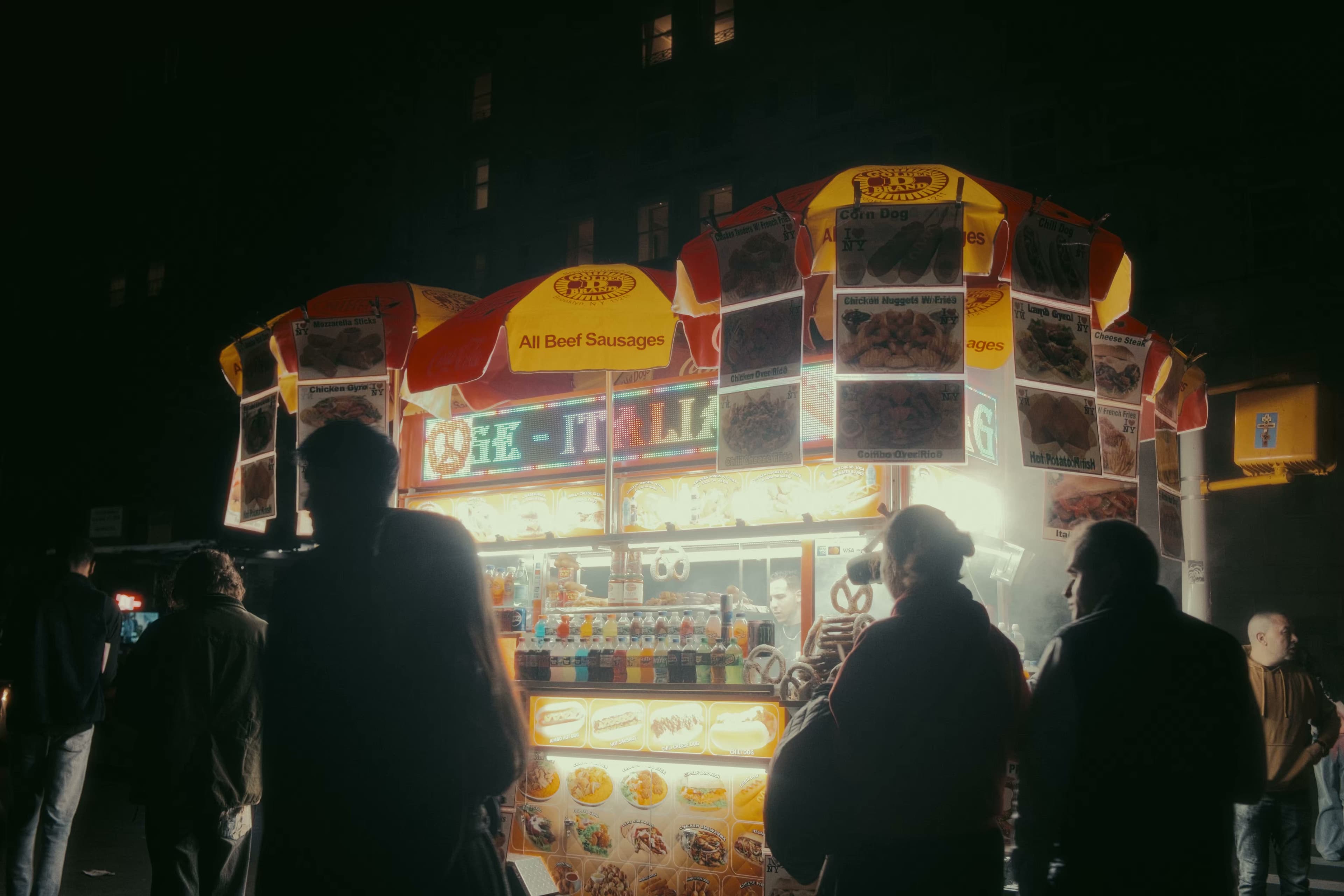 Silhouettes gathered around a glowing NYC street food cart at night.