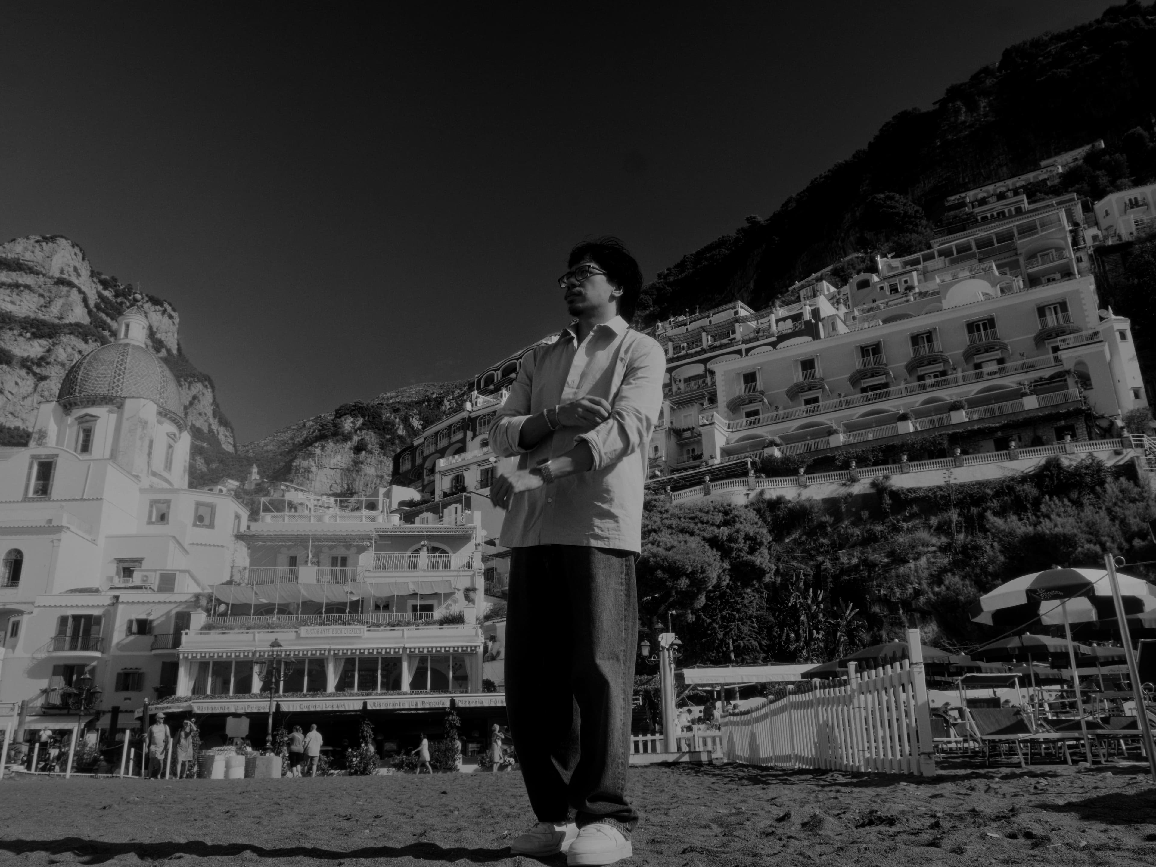 A figure stands on the beach with cliffside buildings rising behind