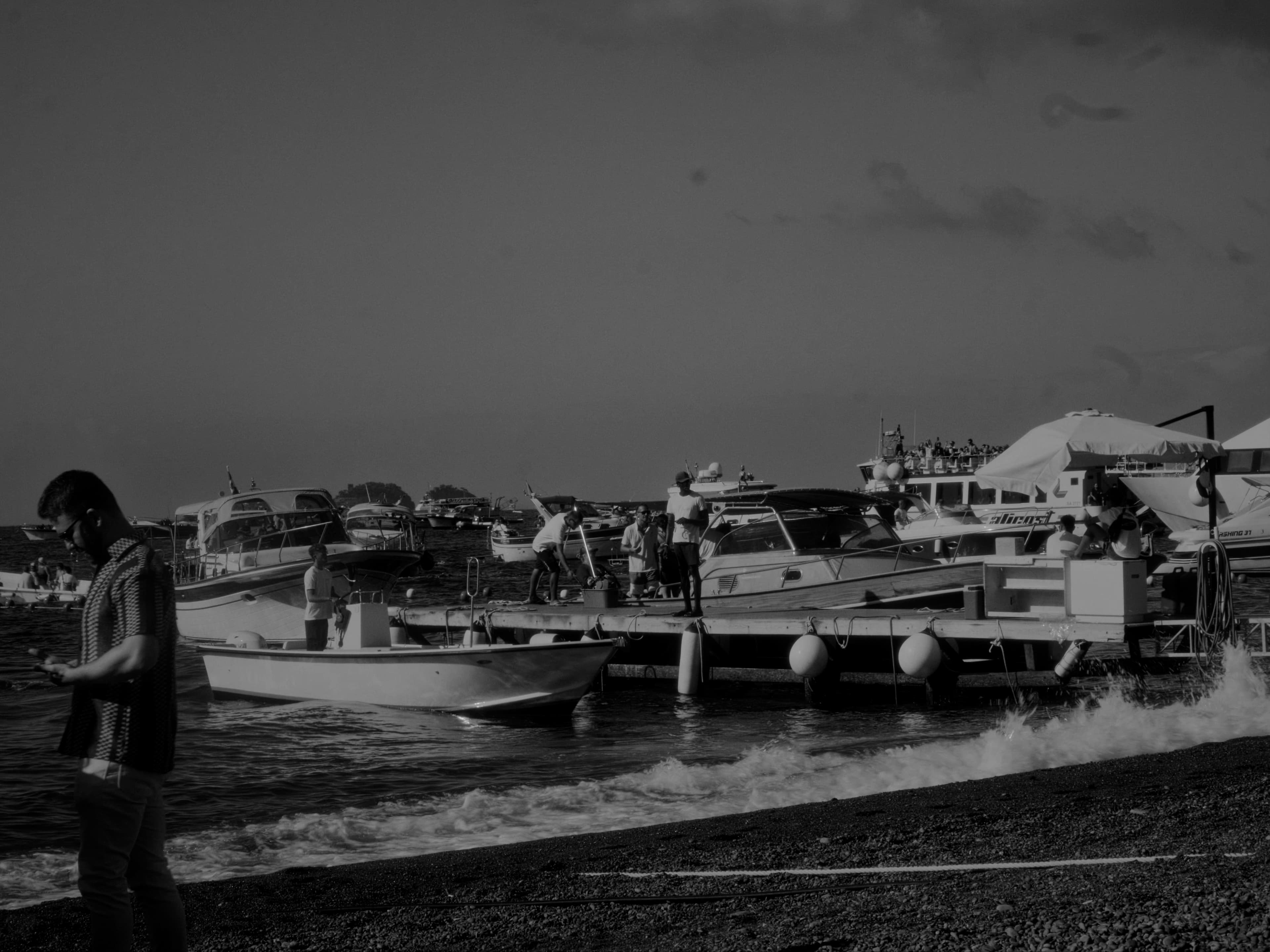 Boats cluster along a shoreline as waves break on a pebbled beach.