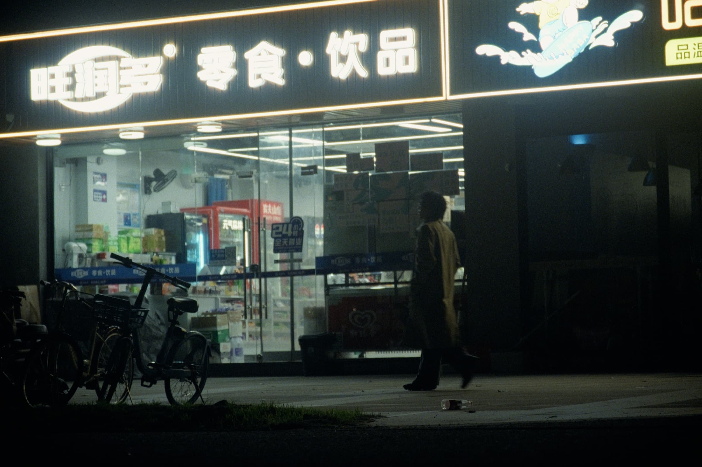 Andrew Amisola in People Who Meet Will Meet Again — A person in a trench coat walking past a brightly lit convenience store at night, bicycles parked in front, Chinese signage glowing above.