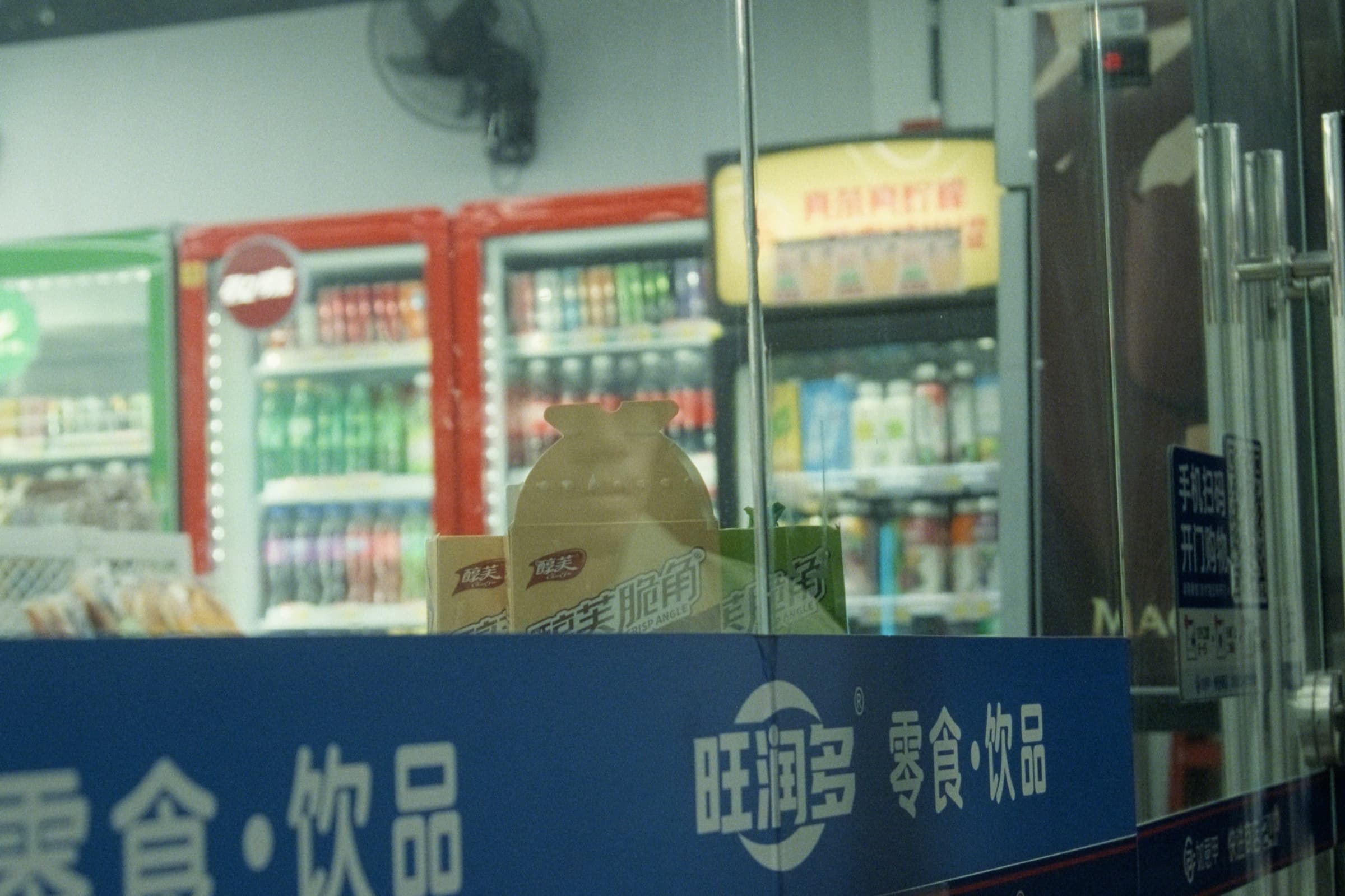 View through a convenience store window showing drink fridges and snack packaging, the store's 旺润多 零食·饮品 logo on the glass.
