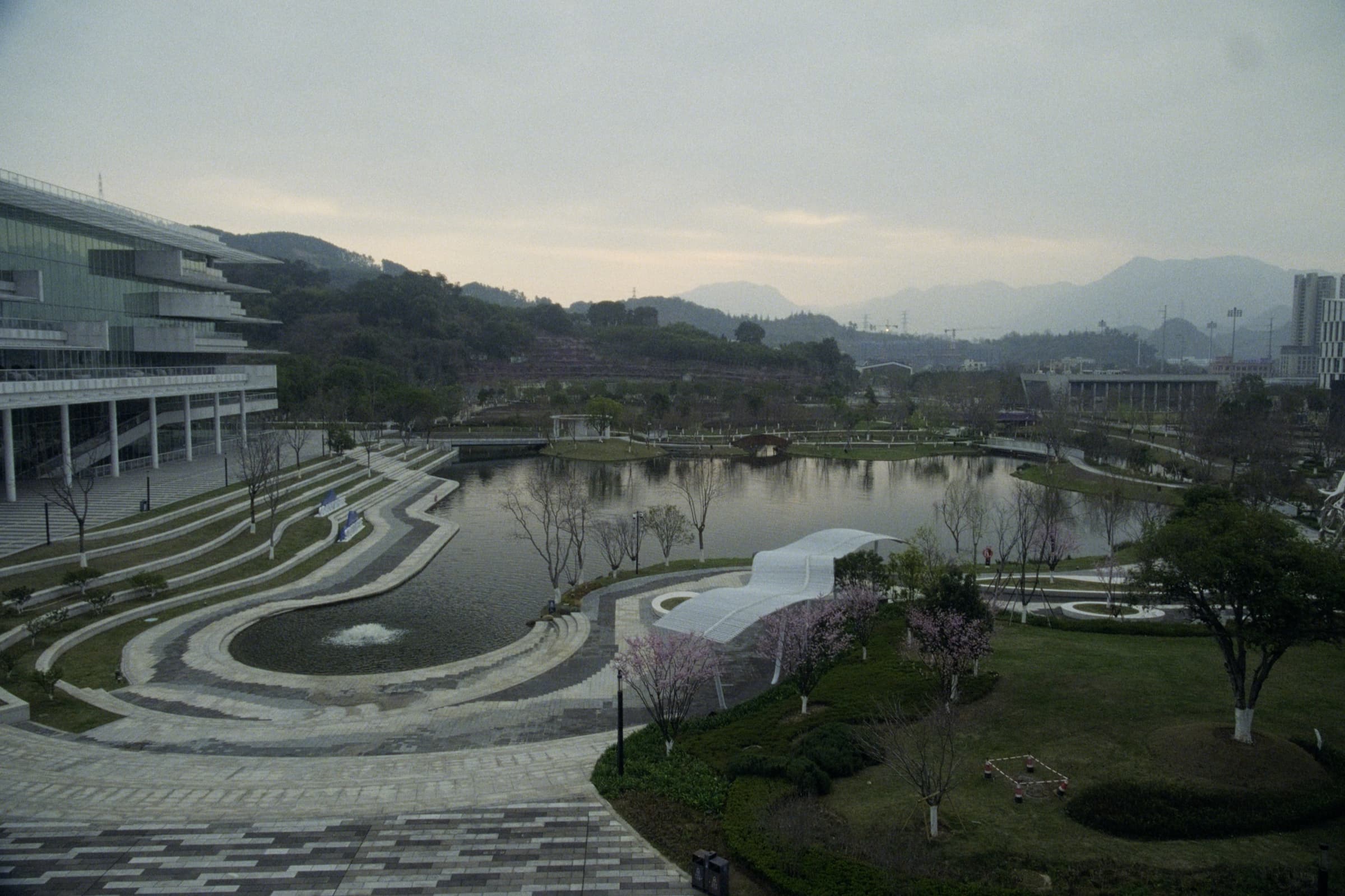 Elevated view of a modern campus park with a curving lake, cherry blossom trees, and misty mountains in the background.