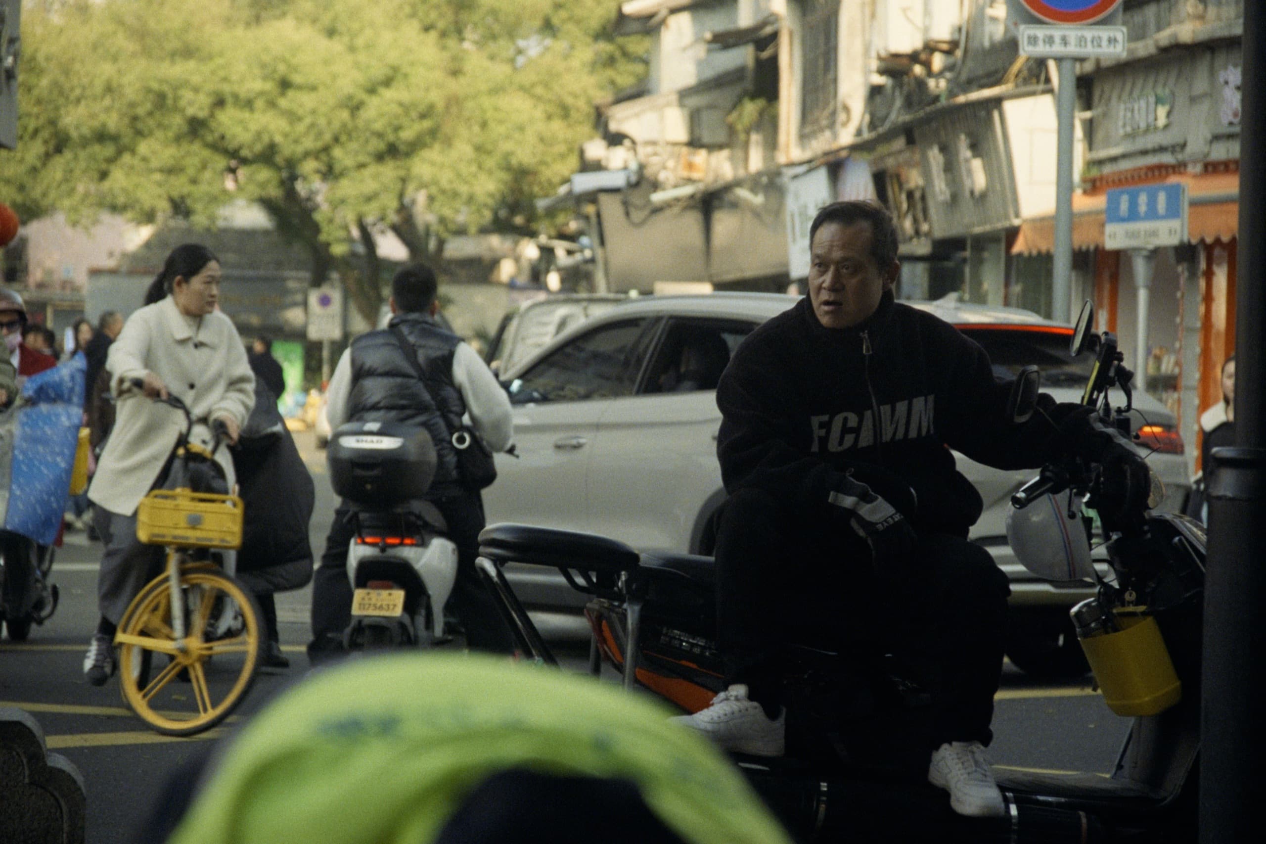 A man on a scooter in a busy Chinese street with pedestrians, bikes, cars, and old storefronts in warm afternoon light.
