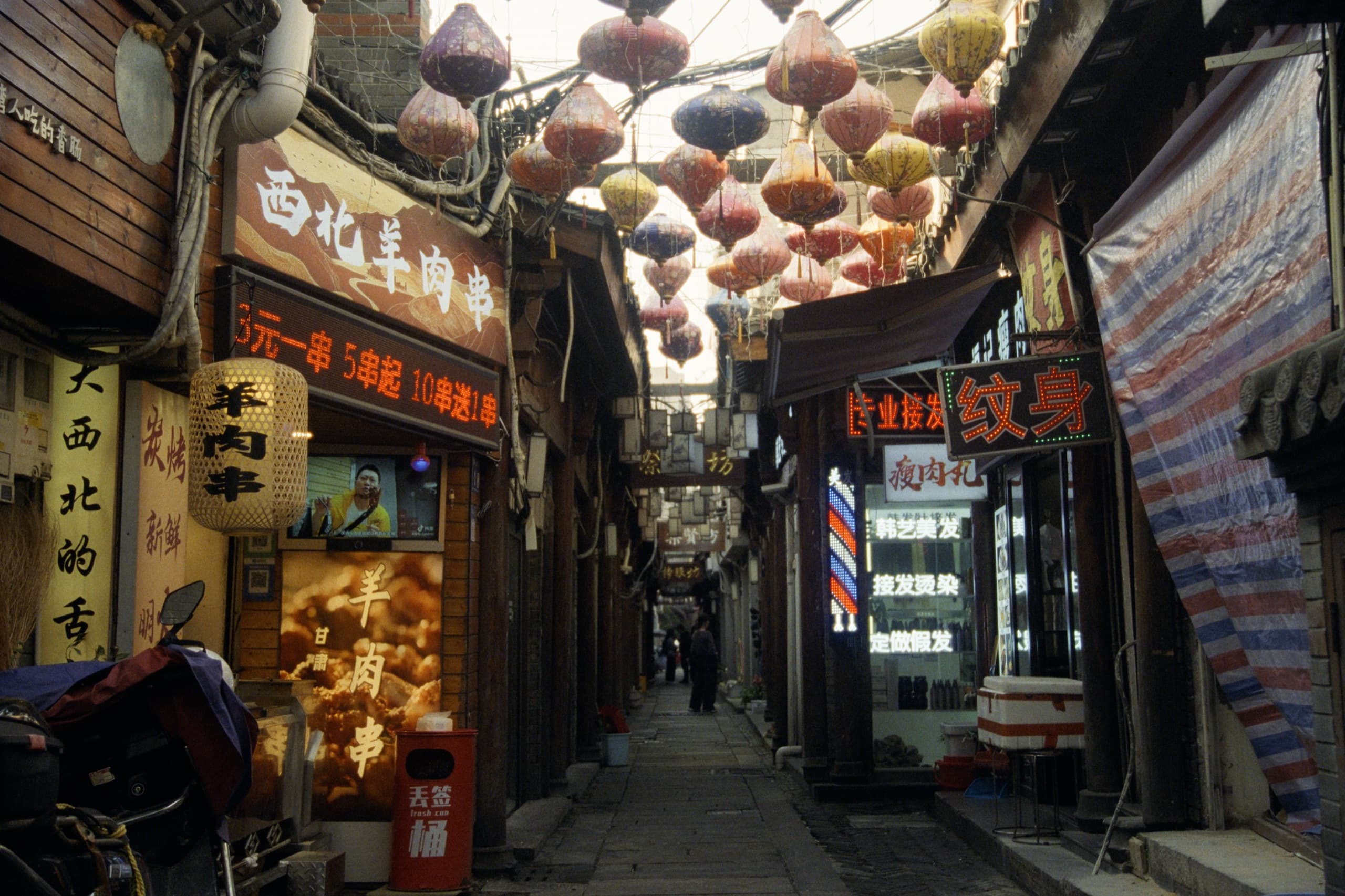 A narrow alley lined with food stalls and shops, colorful lanterns hanging overhead, Chinese signage for lamb skewers and tattoos.