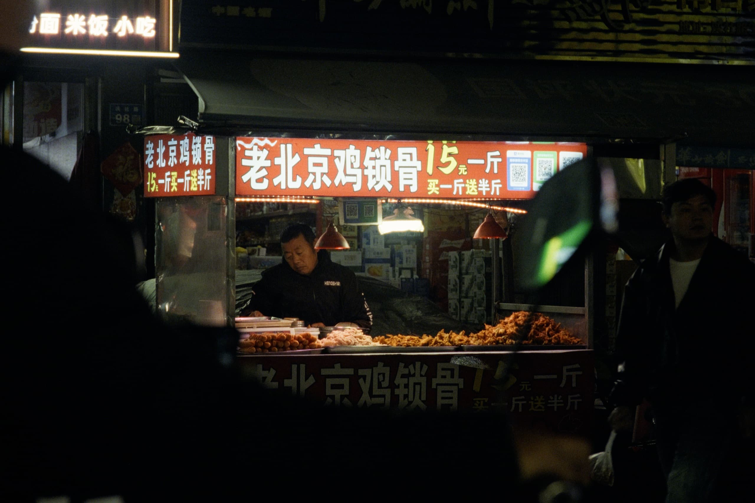A food vendor working behind a stall of fried chicken under red signage reading 老北京鸡锁骨 at night, warm pendant lamps overhead.