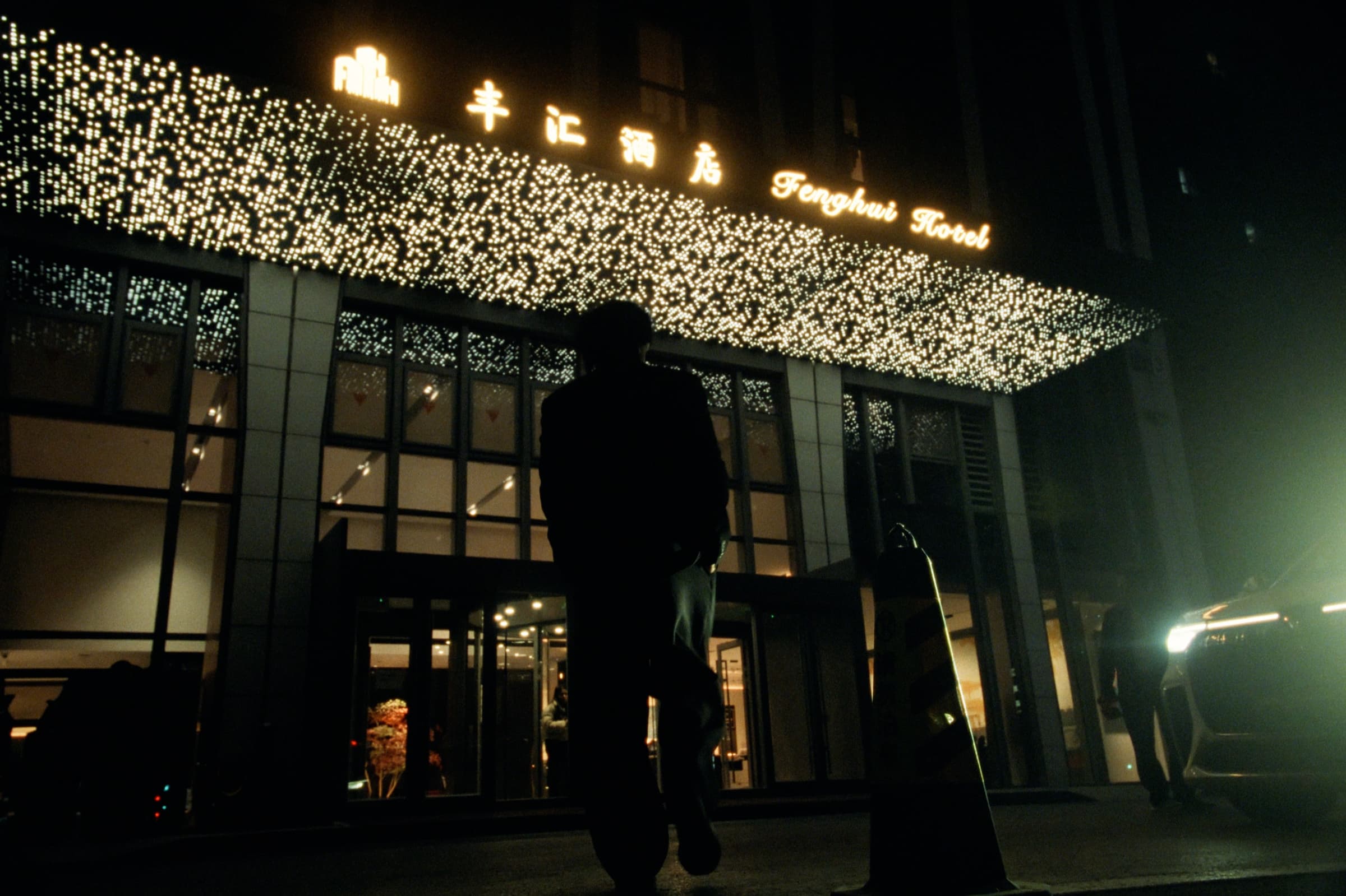 Andrew Amisola in People Who Meet Will Meet Again — A silhouetted figure standing outside the Fenghui Hotel at night, a canopy of fairy lights glittering above the entrance.