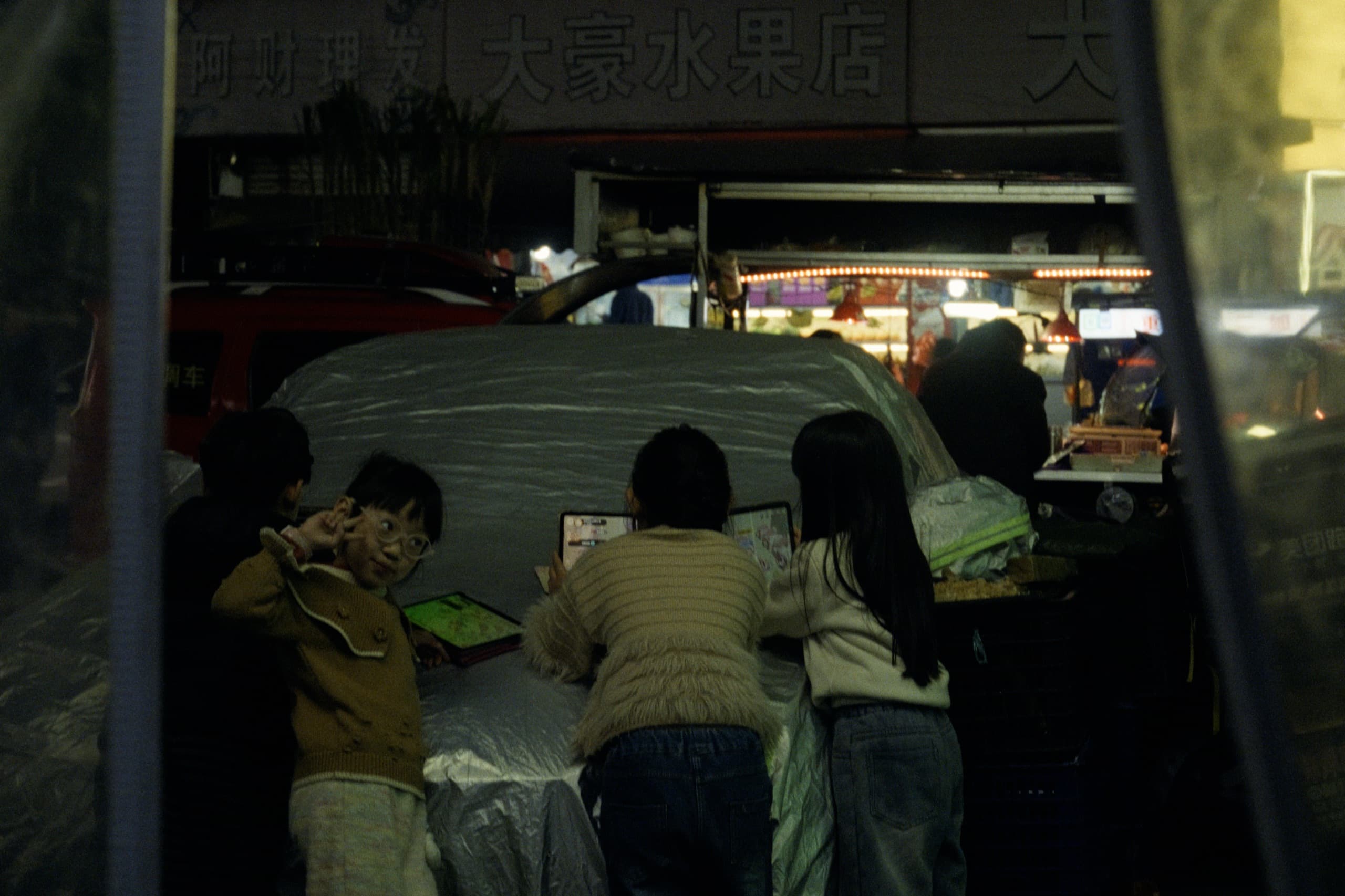 Children huddled around tablets on a covered car hood at night, one boy in glasses looking at the camera, a fruit shop sign behind them.