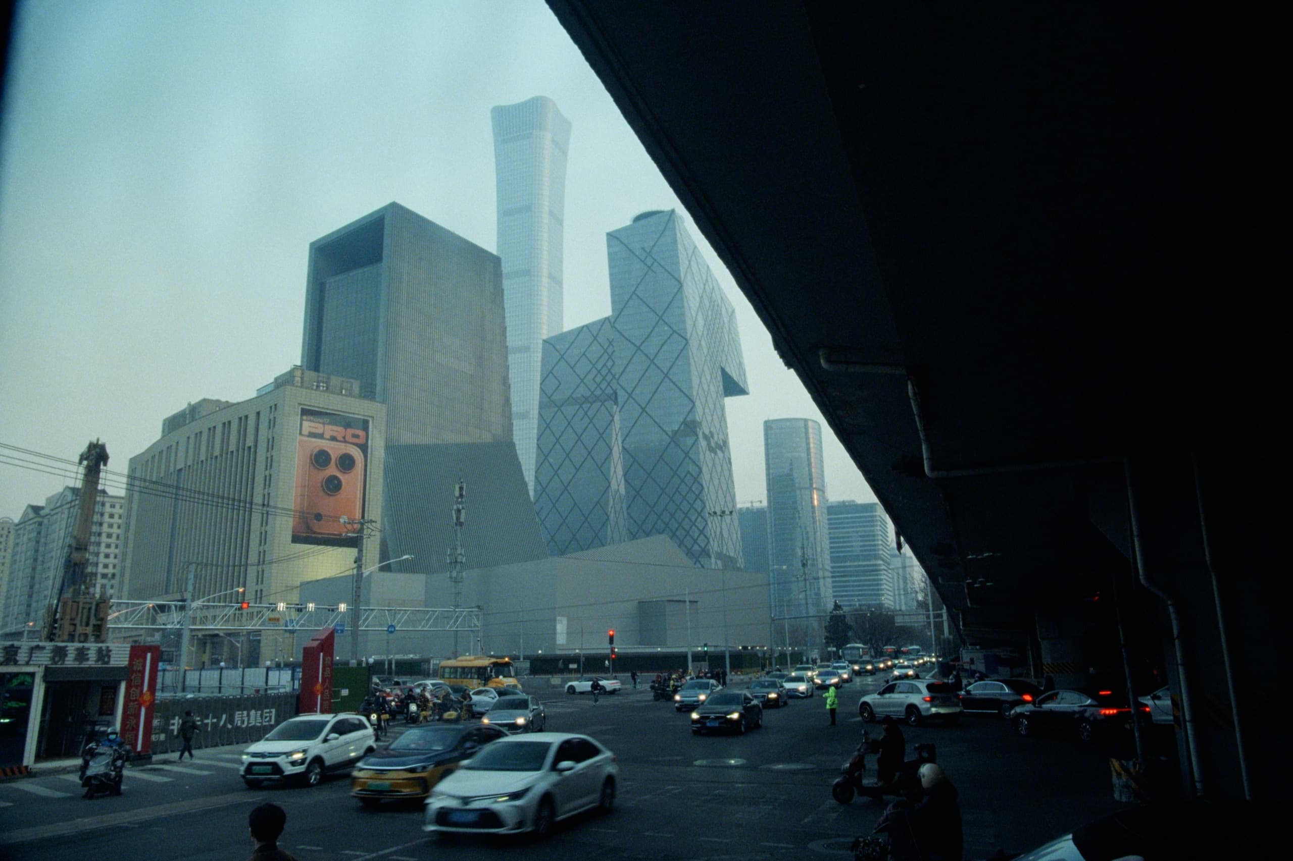 Beijing's CCTV Headquarters and China Zun tower rising through haze, busy traffic below, shot from under a highway overpass.