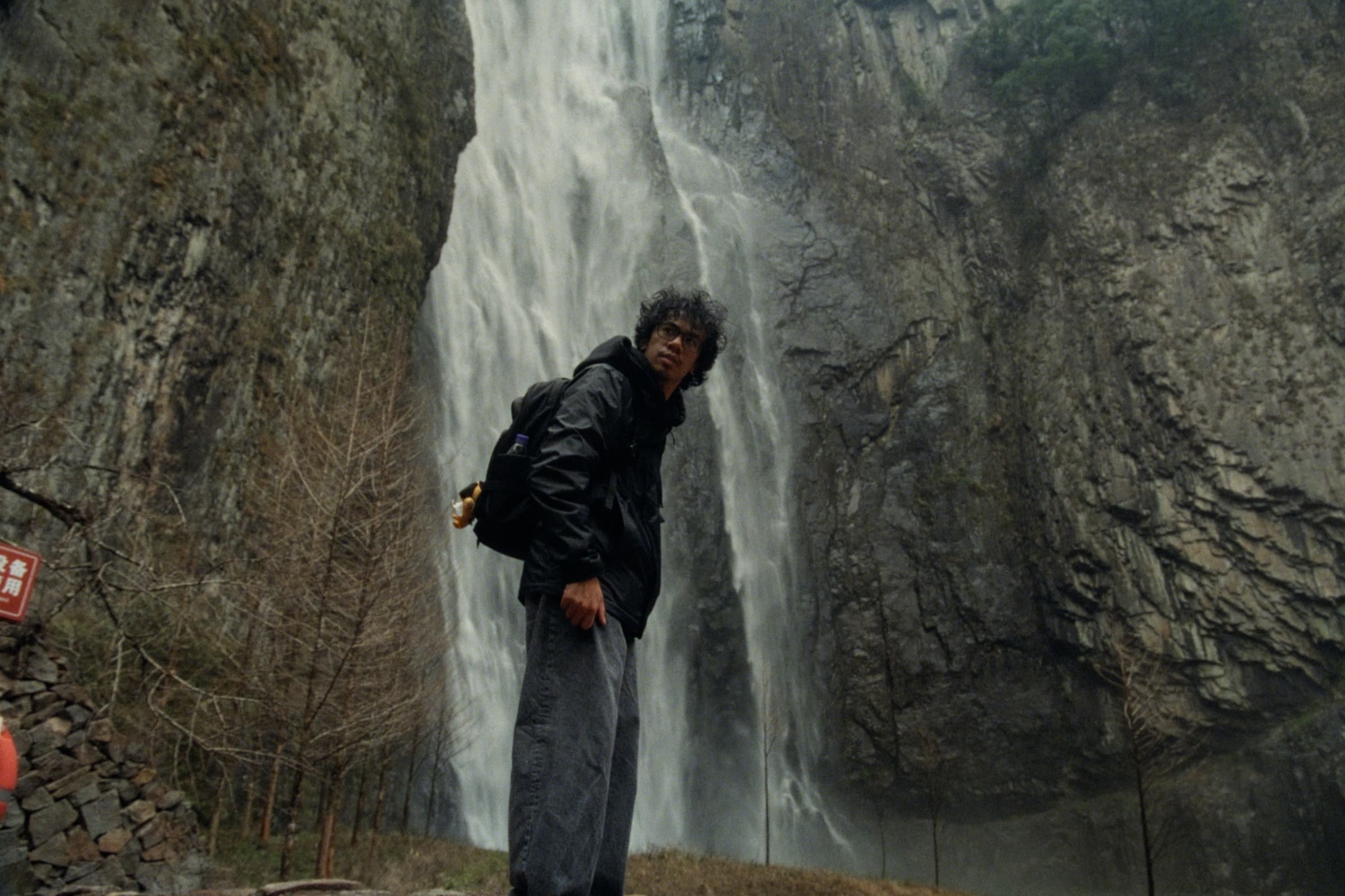 Andrew Amisola in People Who Meet Will Meet Again — Andrew in a black jacket and backpack looking back at the camera with a towering waterfall cascading down a rock face behind him.