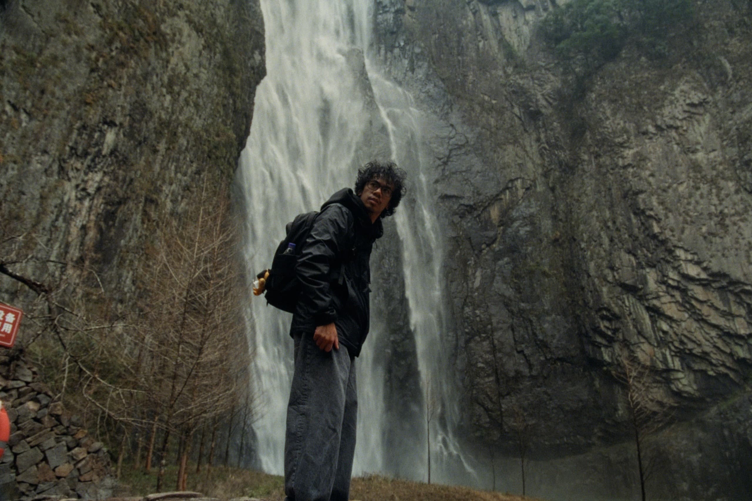 Andrew in a black jacket and backpack looking back at the camera with a towering waterfall cascading down a rock face behind him.
