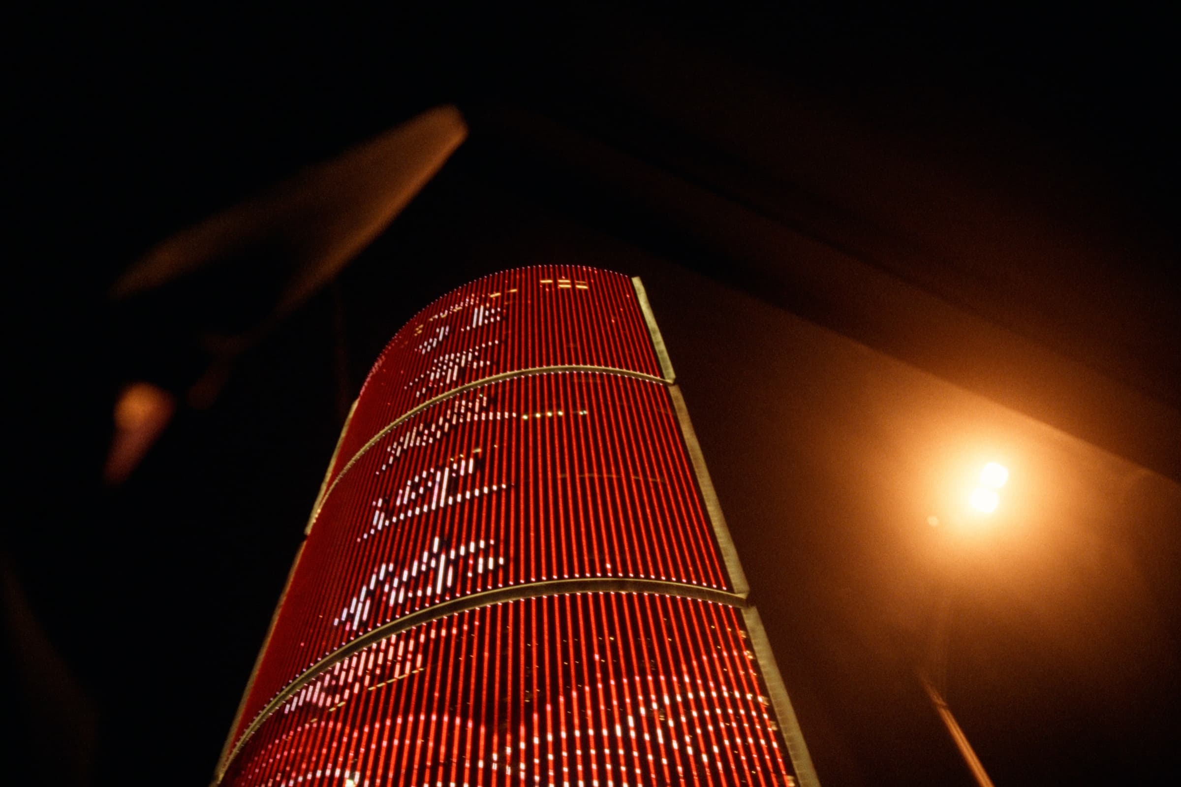 Low-angle night shot of a curved skyscraper lit entirely in red, a street lamp glowing warm to the right.