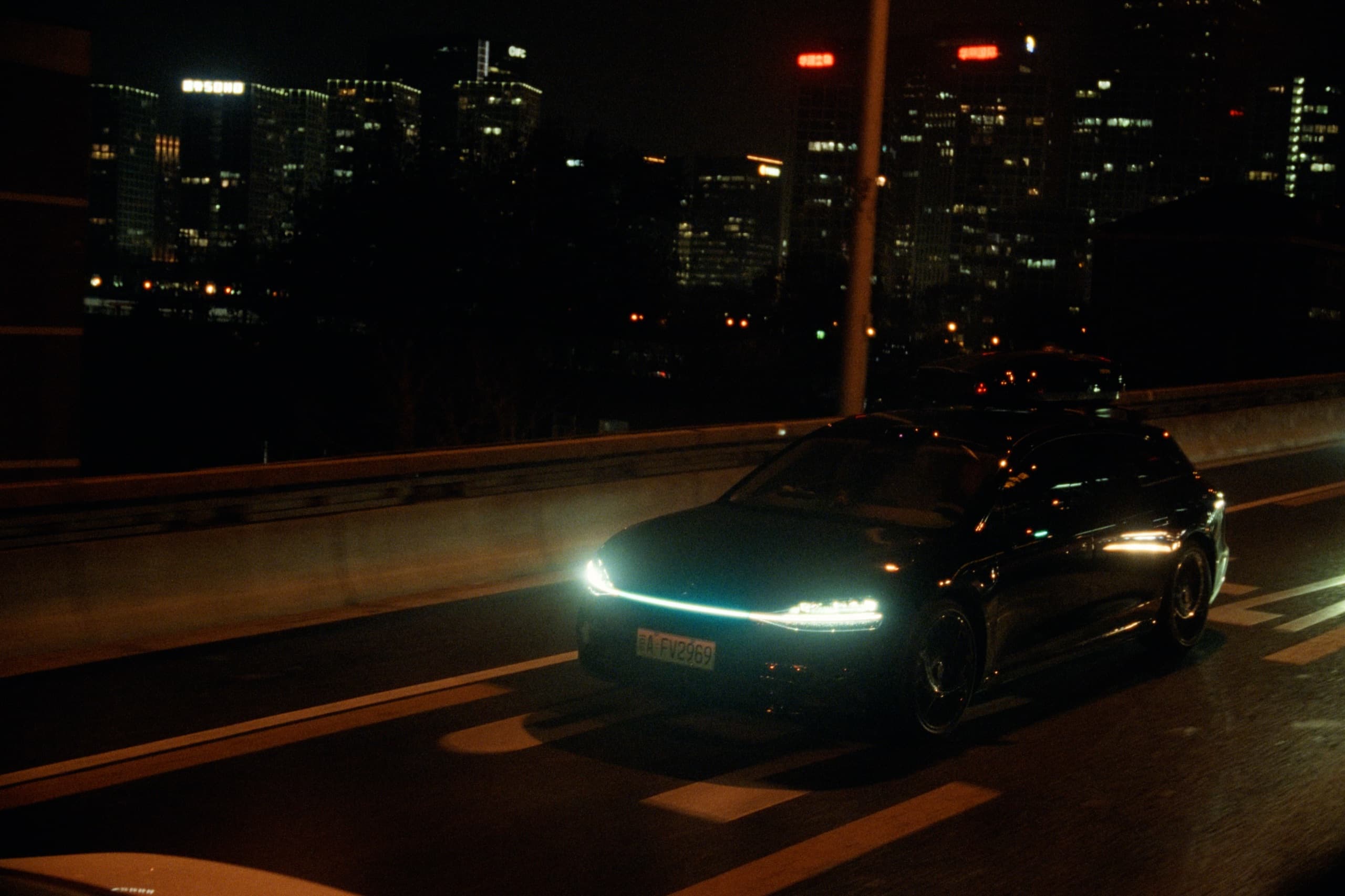 A sleek black car with a glowing LED light bar on a highway at night, a city skyline of illuminated towers in the background.