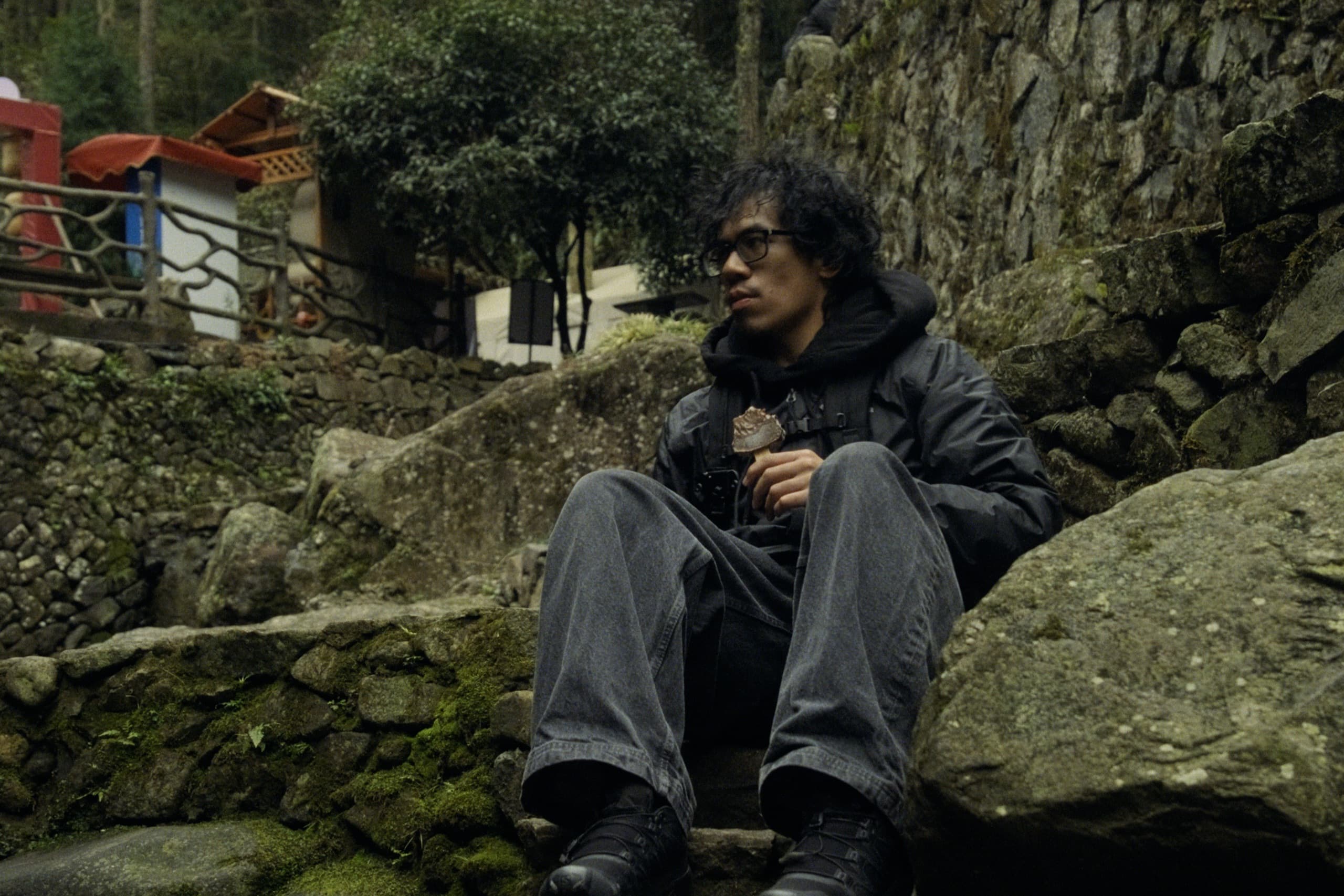 Andrew sitting on mossy rocks eating an ice cream bar, looking off to the side, stone walls and a small pavilion behind him.