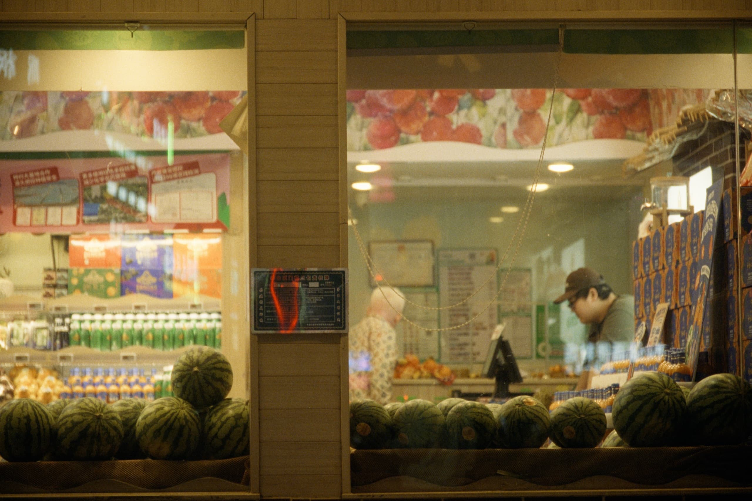 A fruit shop storefront at night with watermelons lined up on the ledge outside and shoppers visible through the glass windows.