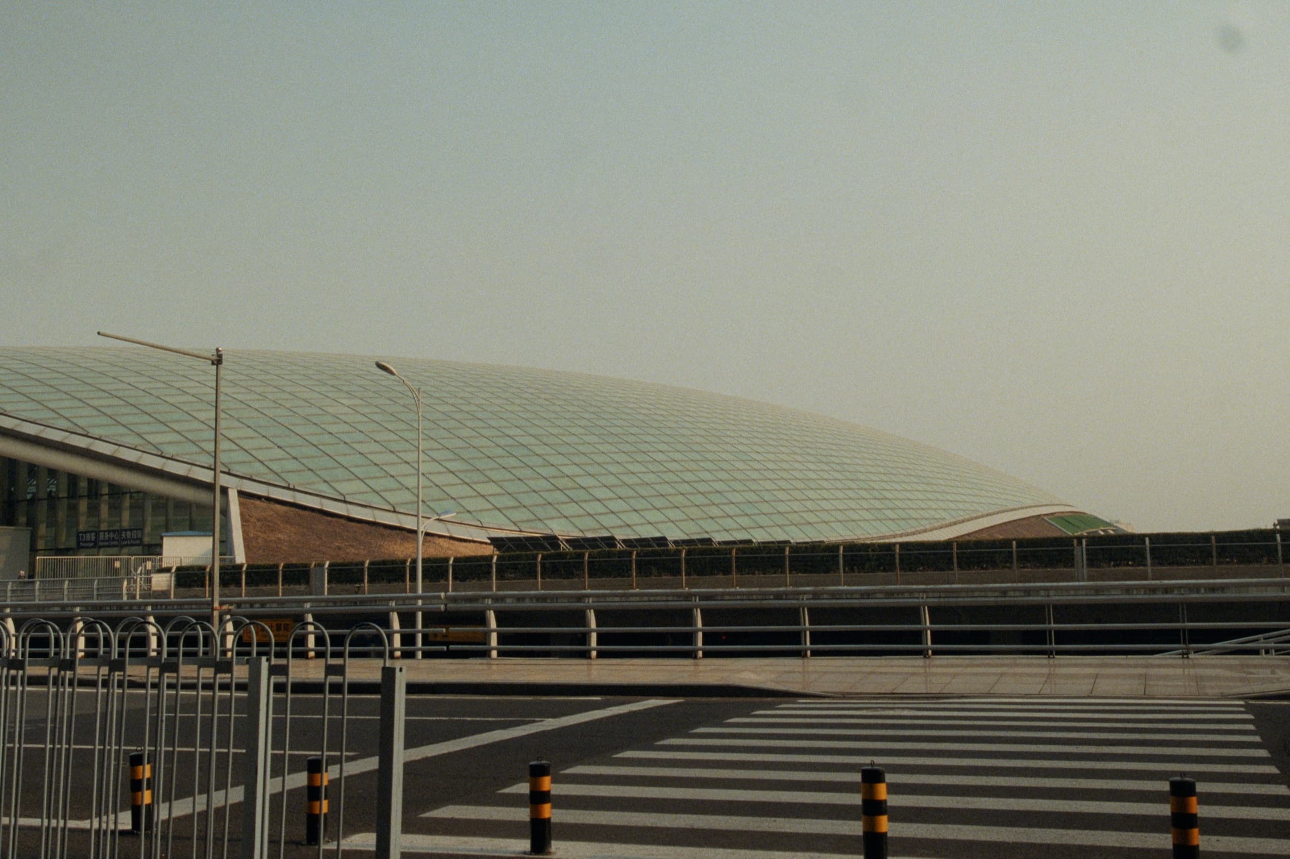 The sweeping glass and steel roof of Beijing Capital International Airport seen from the road, crosswalk and bollards in the foreground.