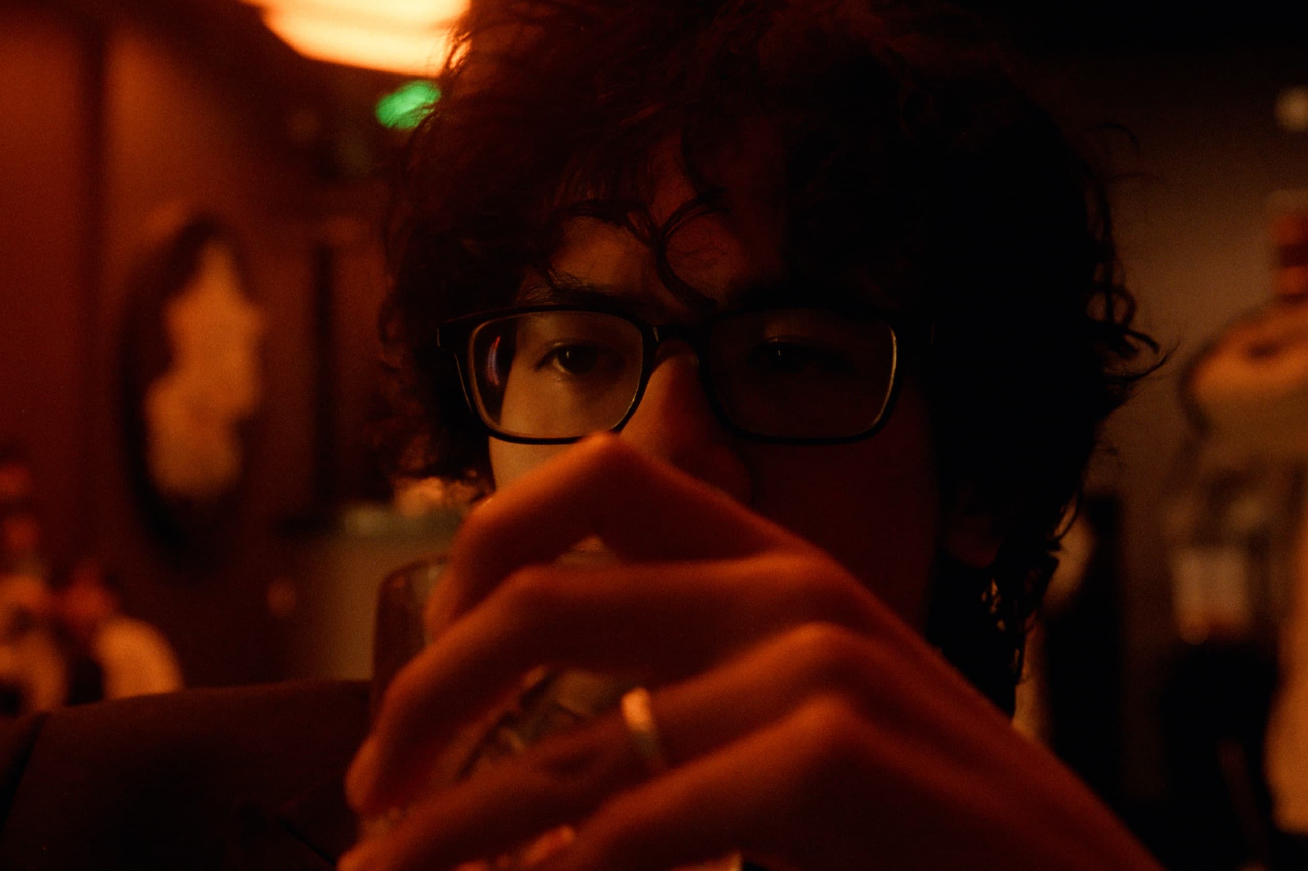 Extreme close-up of Andrew in a warmly lit bar, hands clasped near his chin, glasses catching amber light, rings on his fingers.