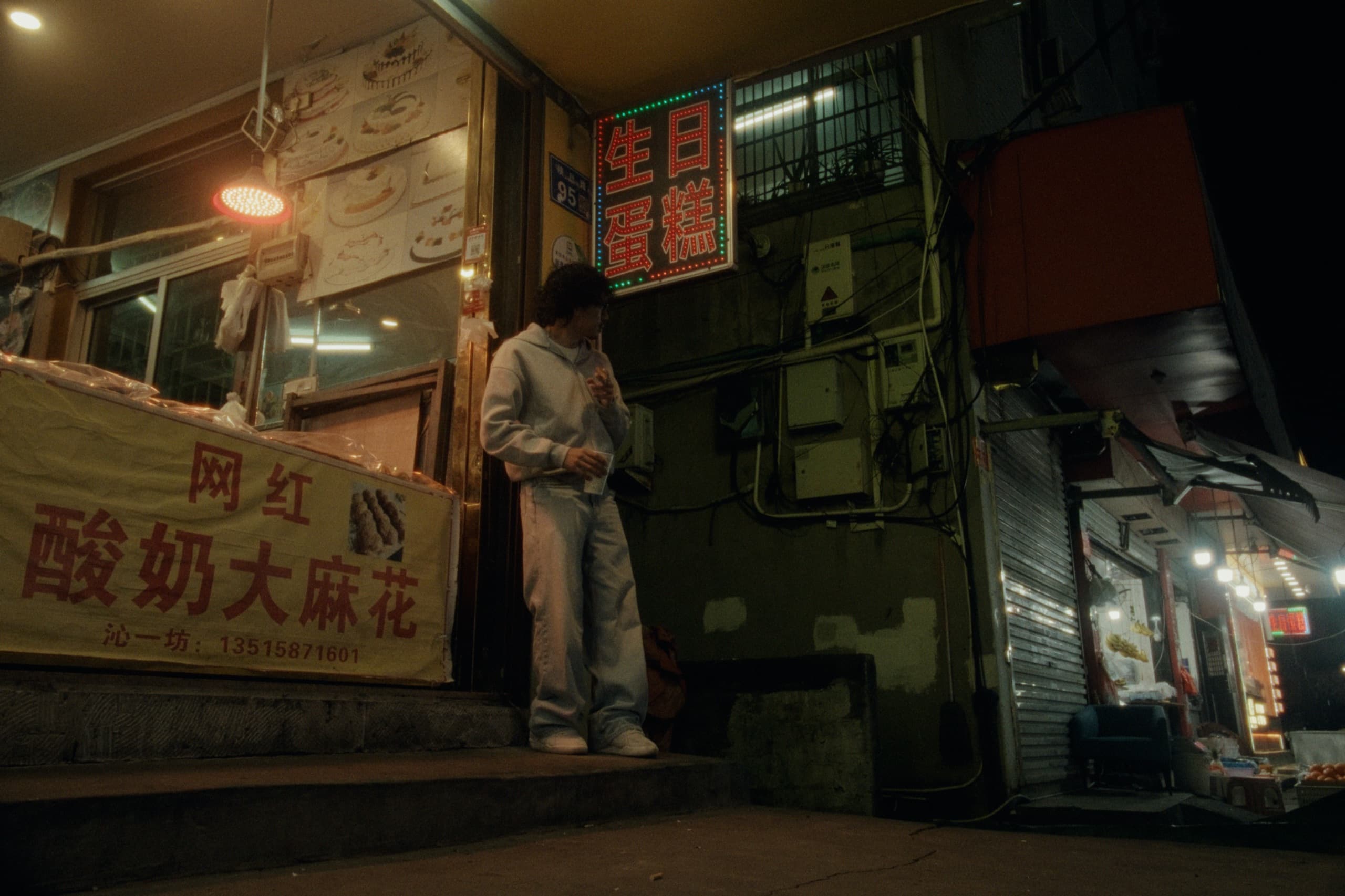 Andrew standing in a gray hoodie outside a bakery at night, an LED sign for 生日蛋糕 above and a banner for 网红酸奶大麻花 beside him.