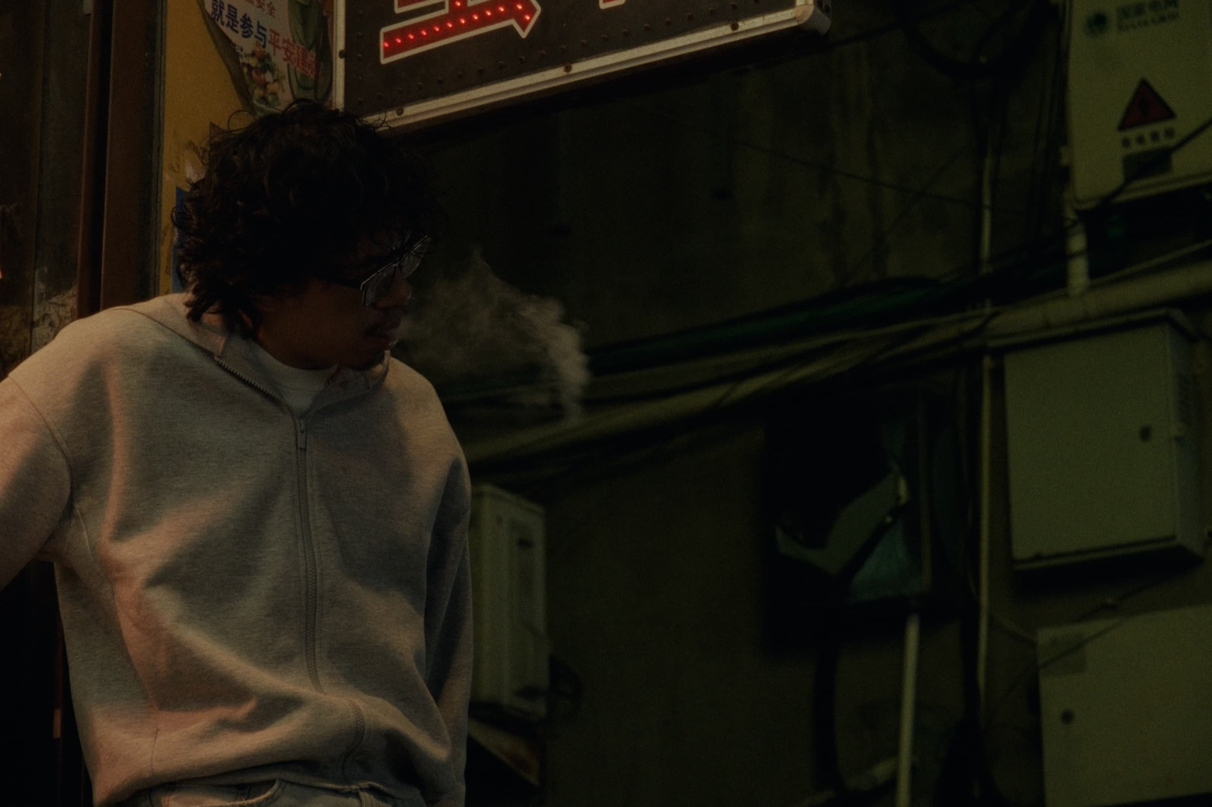 Andrew Amisola in People Who Meet Will Meet Again — Close-up of Andrew in a light gray zip-up sweatshirt exhaling visible breath into the cold night air, an LED arrow sign glowing behind him.