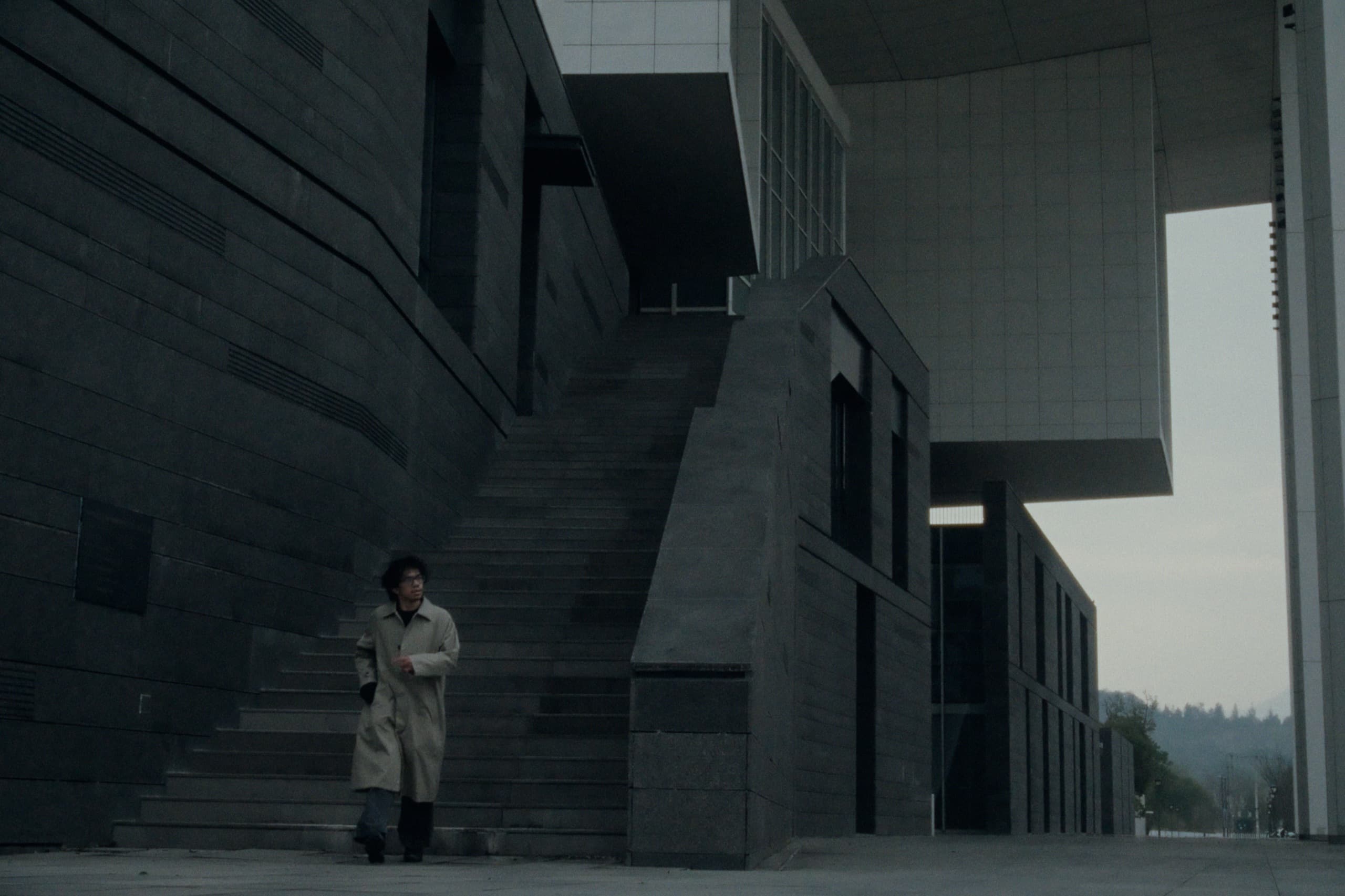 Andrew in a beige trench coat dwarfed by a massive brutalist building with angular staircases and dark stone walls.
