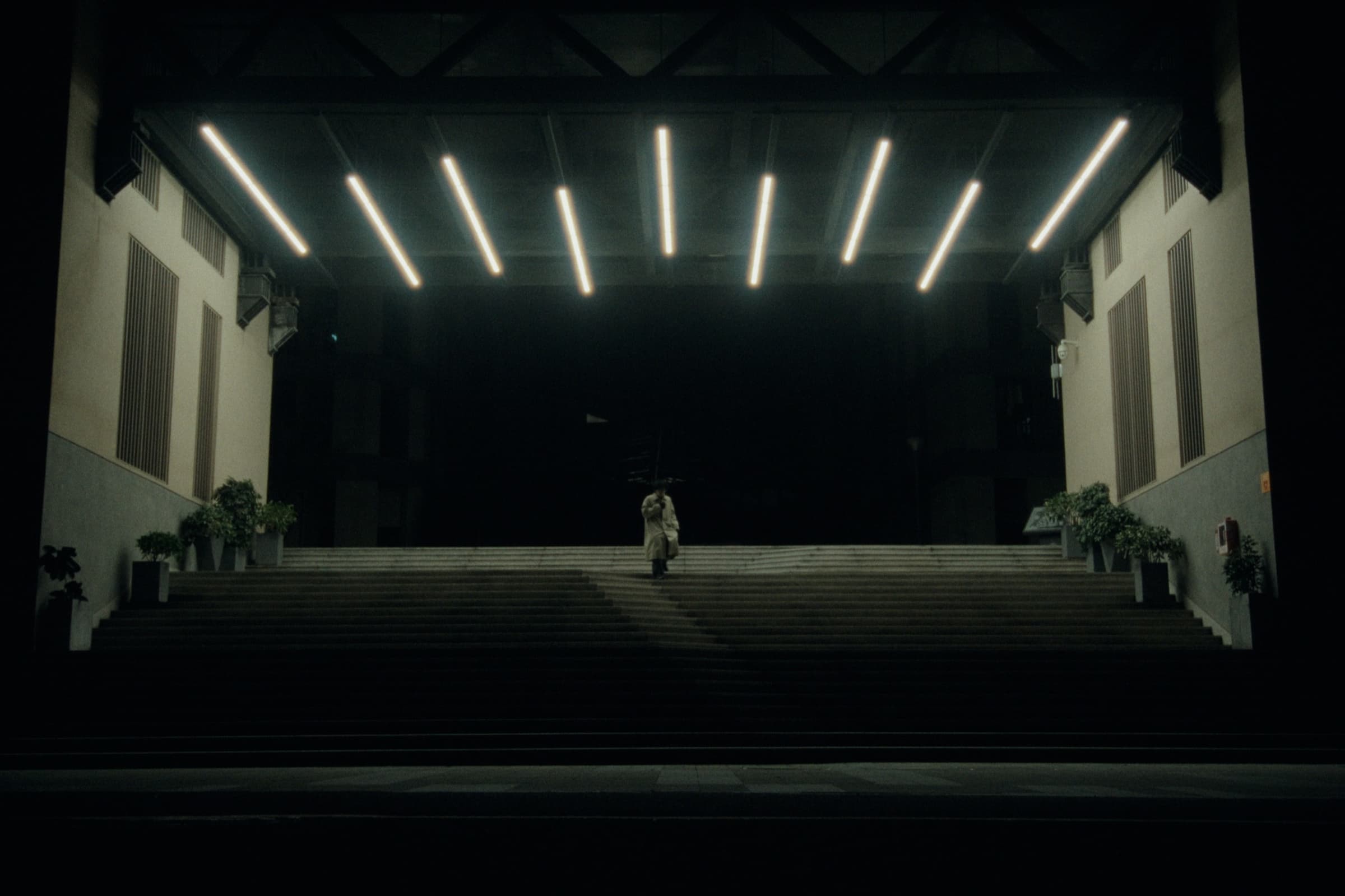 Andrew Amisola in People Who Meet Will Meet Again — A lone figure in a trench coat ascending wide concrete steps under parallel fluorescent tube lights, surrounded by total darkness.