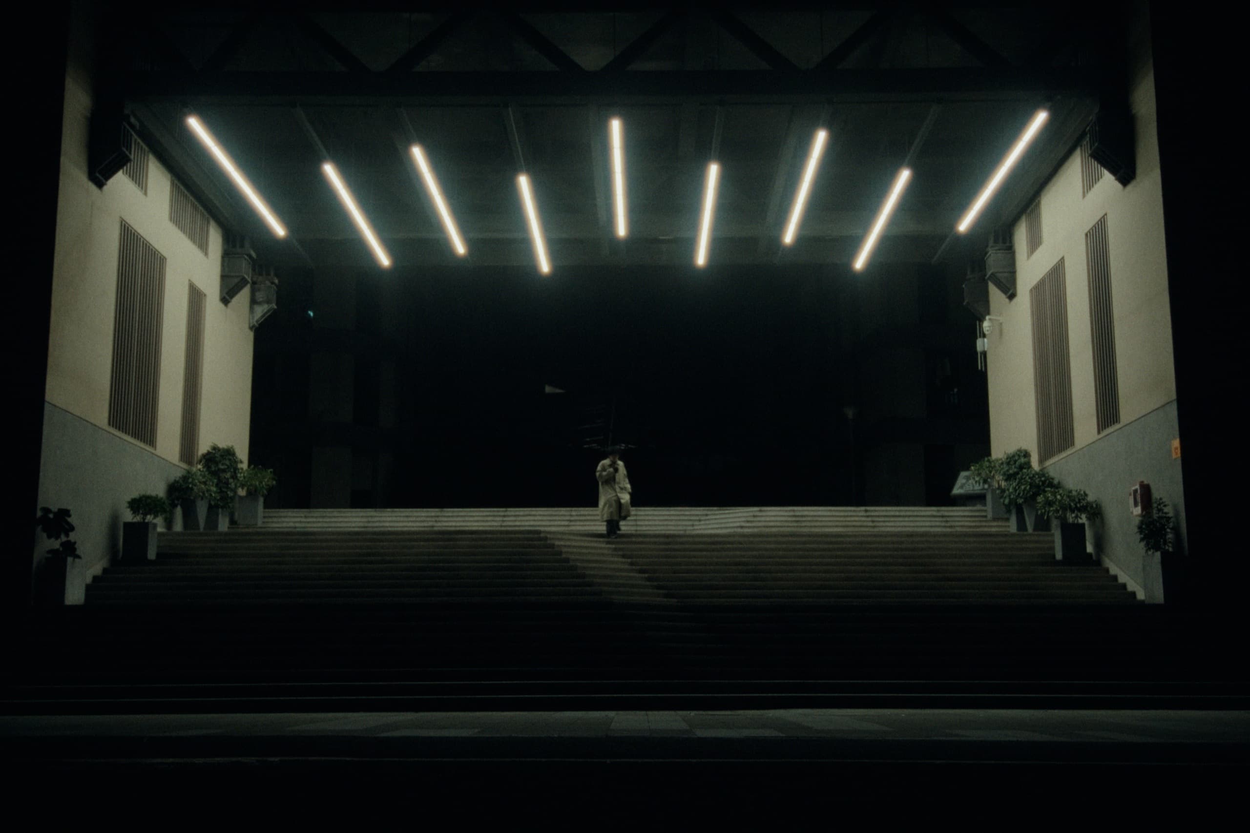 A lone figure in a trench coat ascending wide concrete steps under parallel fluorescent tube lights, surrounded by total darkness.