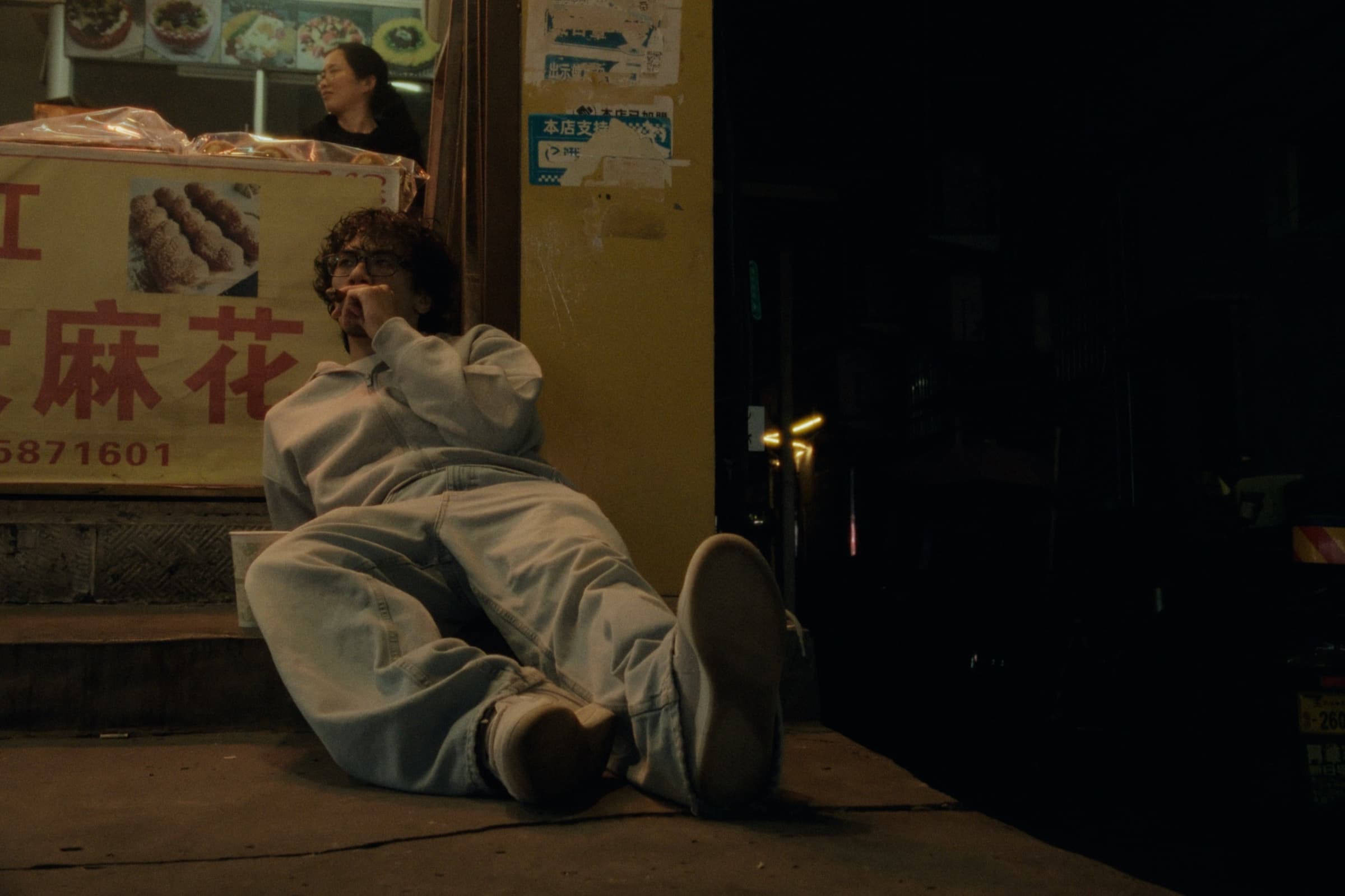 Andrew Amisola in People Who Meet Will Meet Again — Andrew lounging on the sidewalk outside a late-night food stall, legs stretched out, a vendor visible behind him.