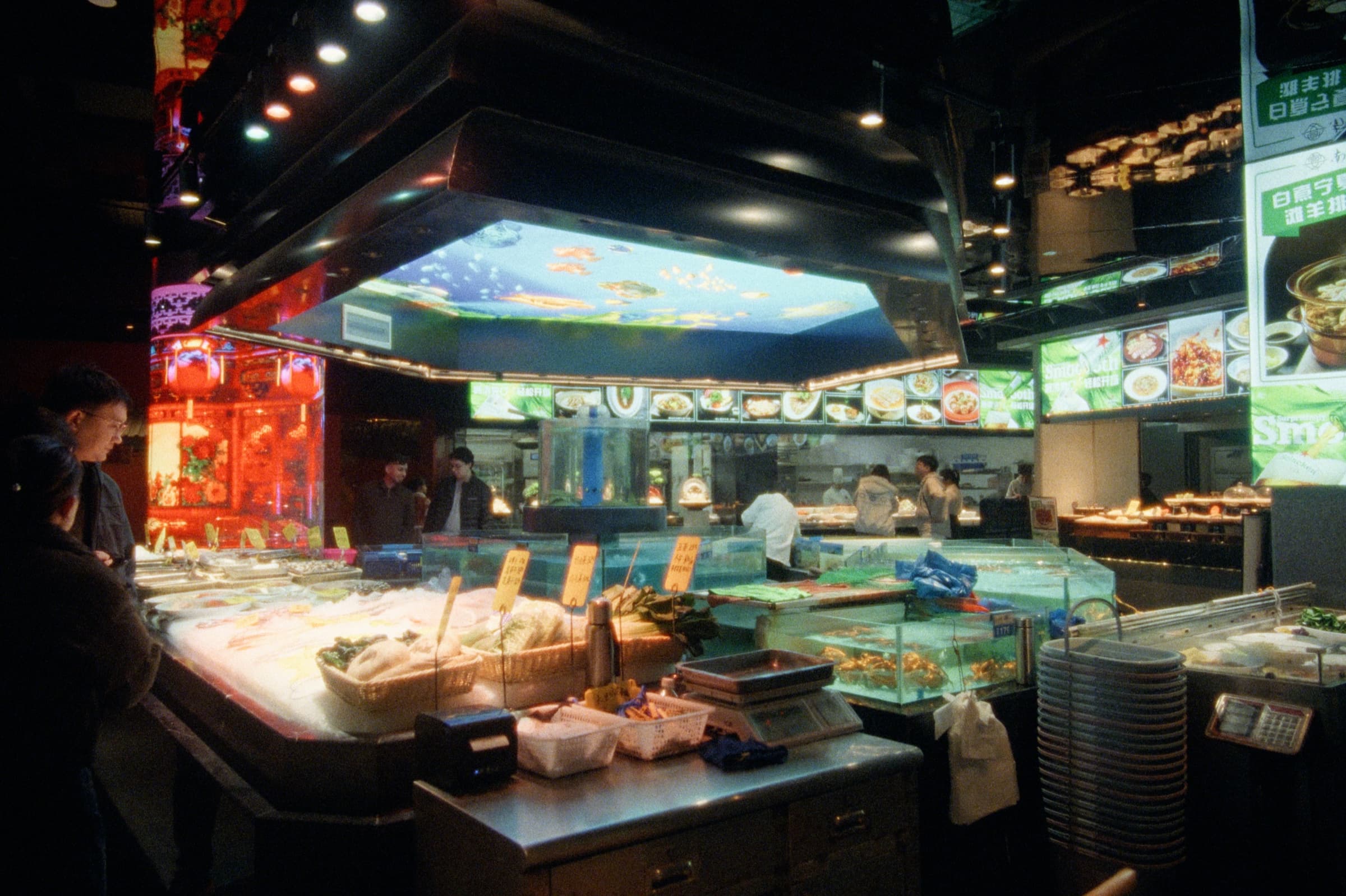 A bustling seafood restaurant interior with glass tanks of live fish, illuminated menu boards, red lanterns, and an aquarium panel glowing on the ceiling.