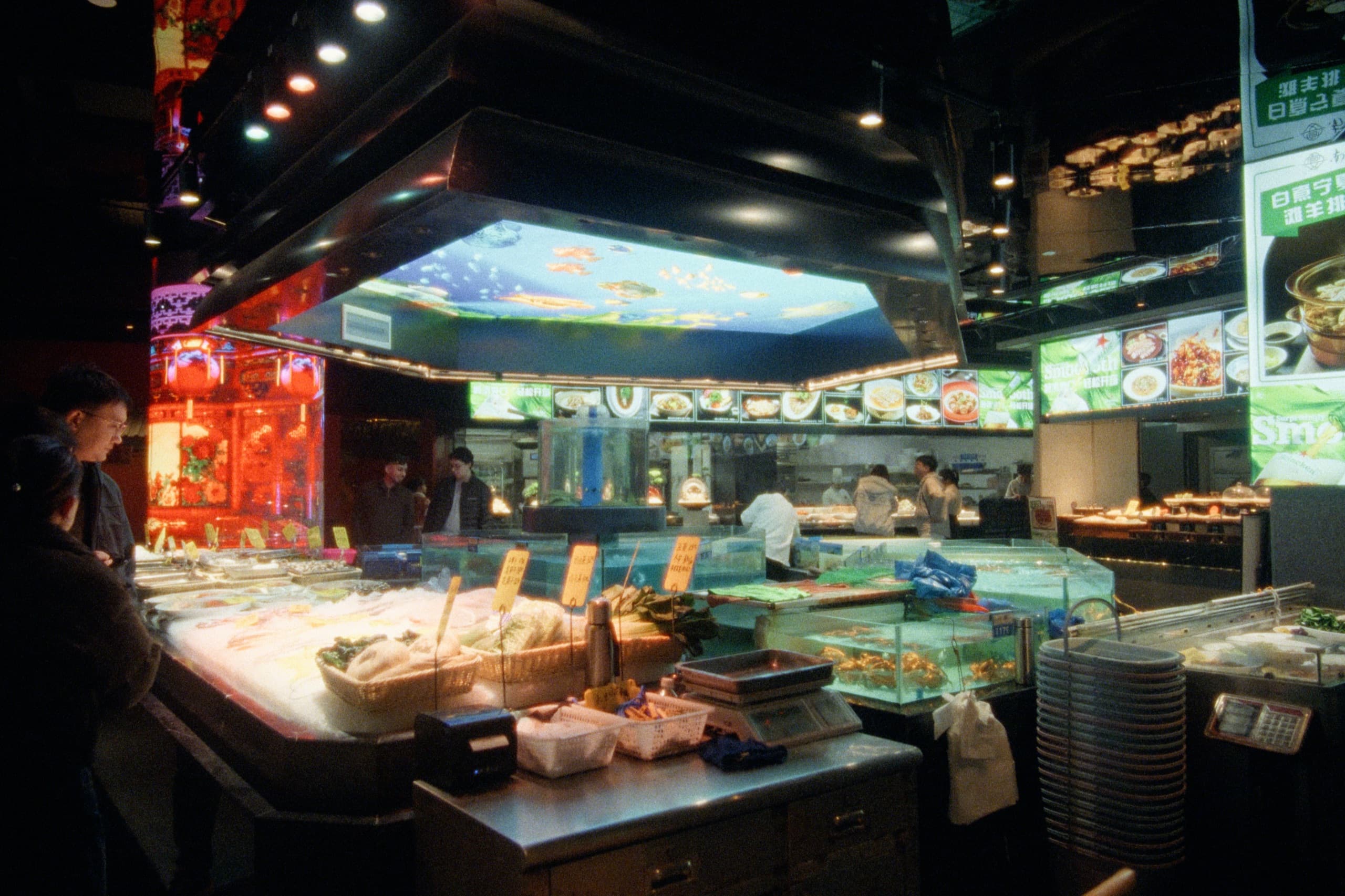 A bustling seafood restaurant interior with glass tanks of live fish, illuminated menu boards, red lanterns, and an aquarium panel glowing on the ceiling.