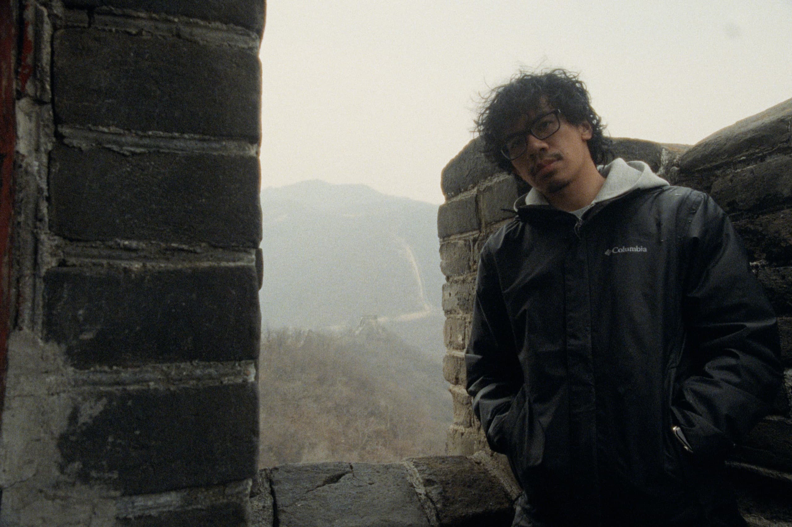 Andrew leaning against the Great Wall in a Columbia jacket, the Wall's ridgeline snaking into hazy mountains through an opening between brick pillars.