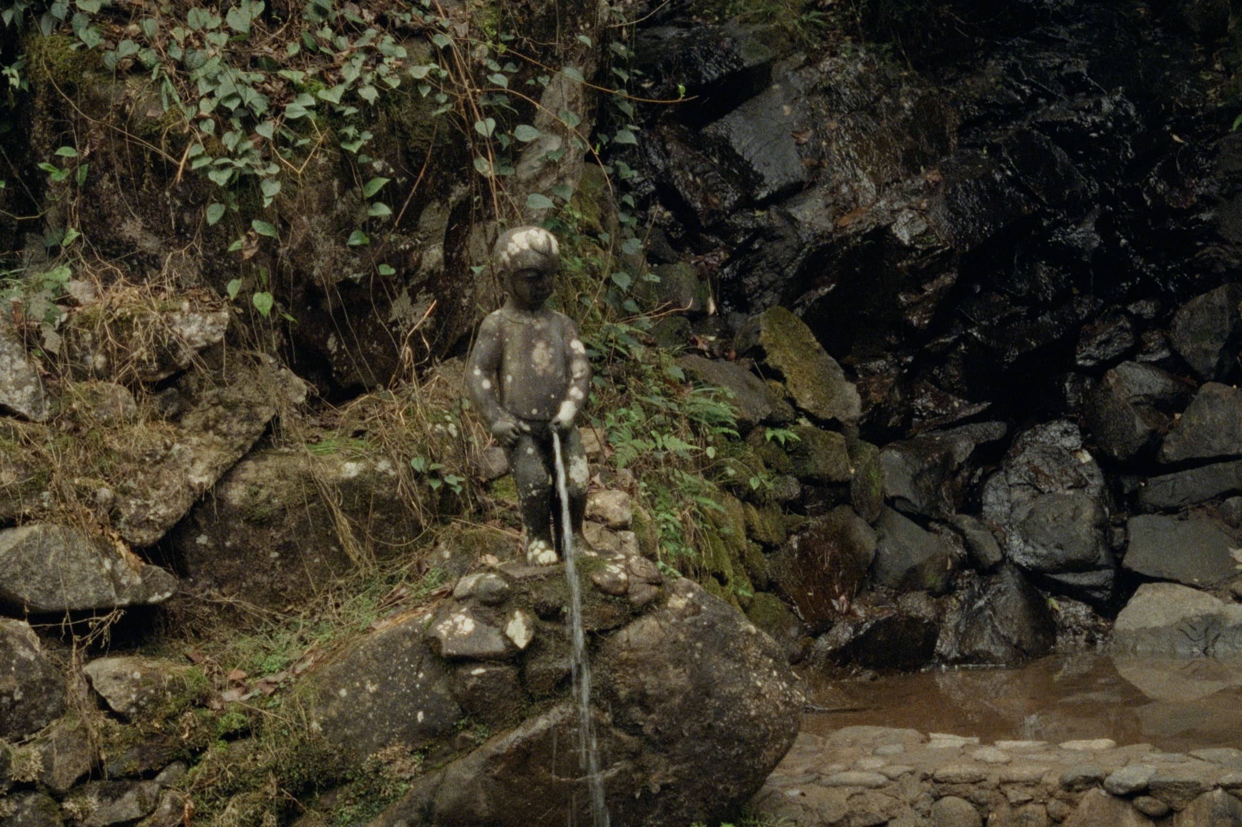 A small weathered stone fountain statue of a peeing boy on mossy rocks, water trickling down into a stream below.
