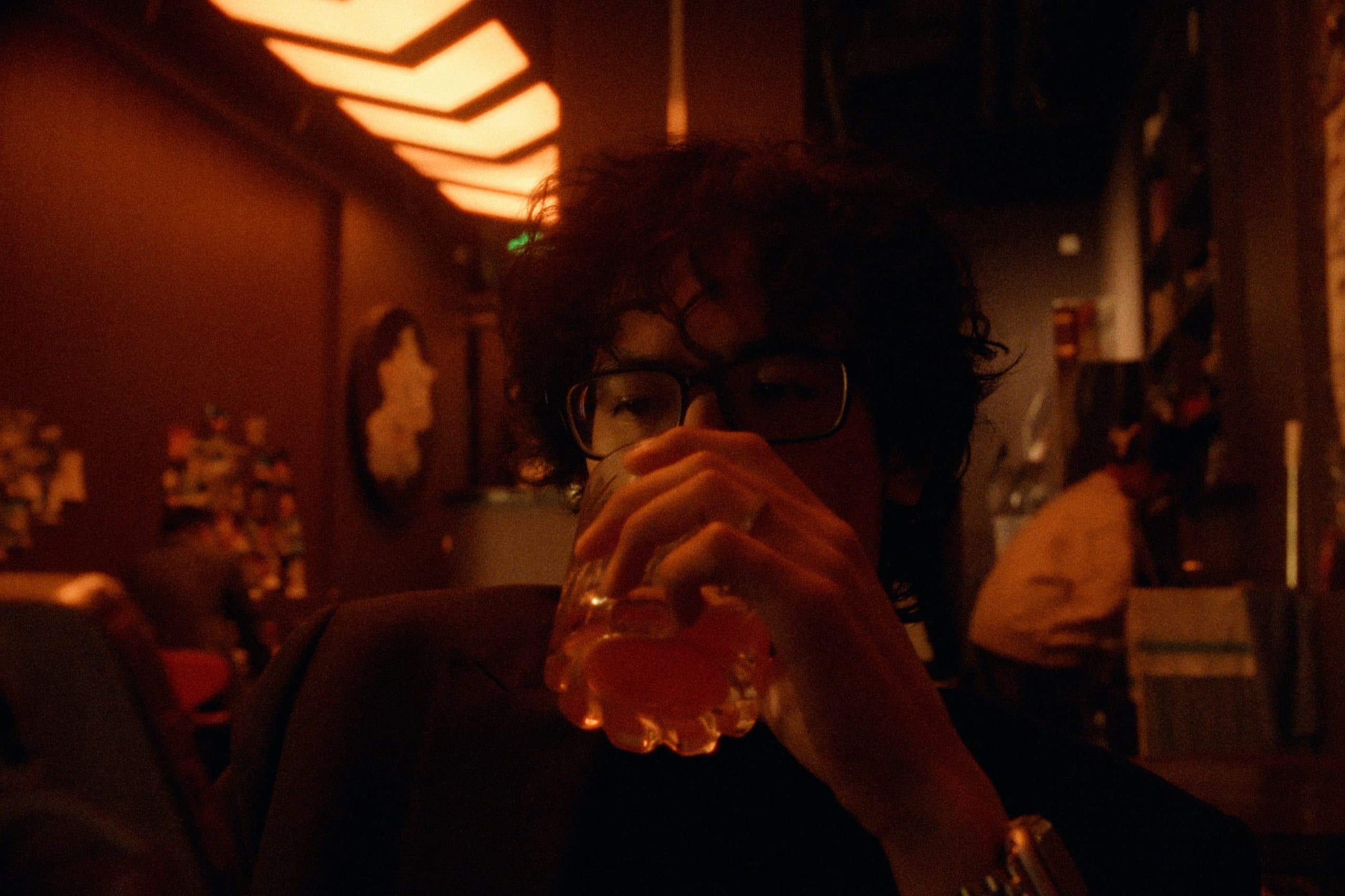 Andrew drinking from a glass in a bar bathed in deep amber light, a chevron-patterned lamp glowing above him.